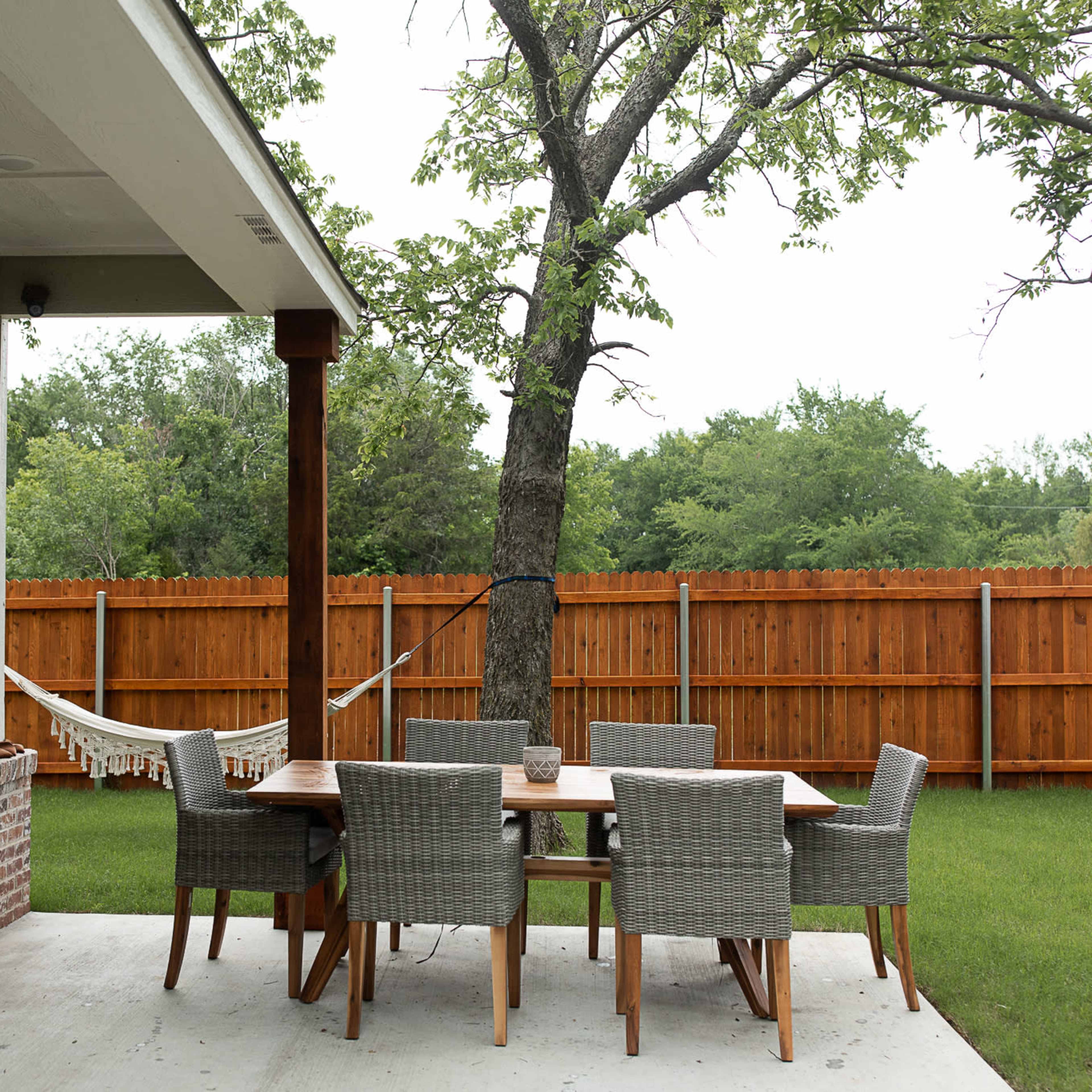 A patio area features a wooden dining table with six chairs, a grill, and a hammock hanging between two trees, surrounded by a fenced yard.