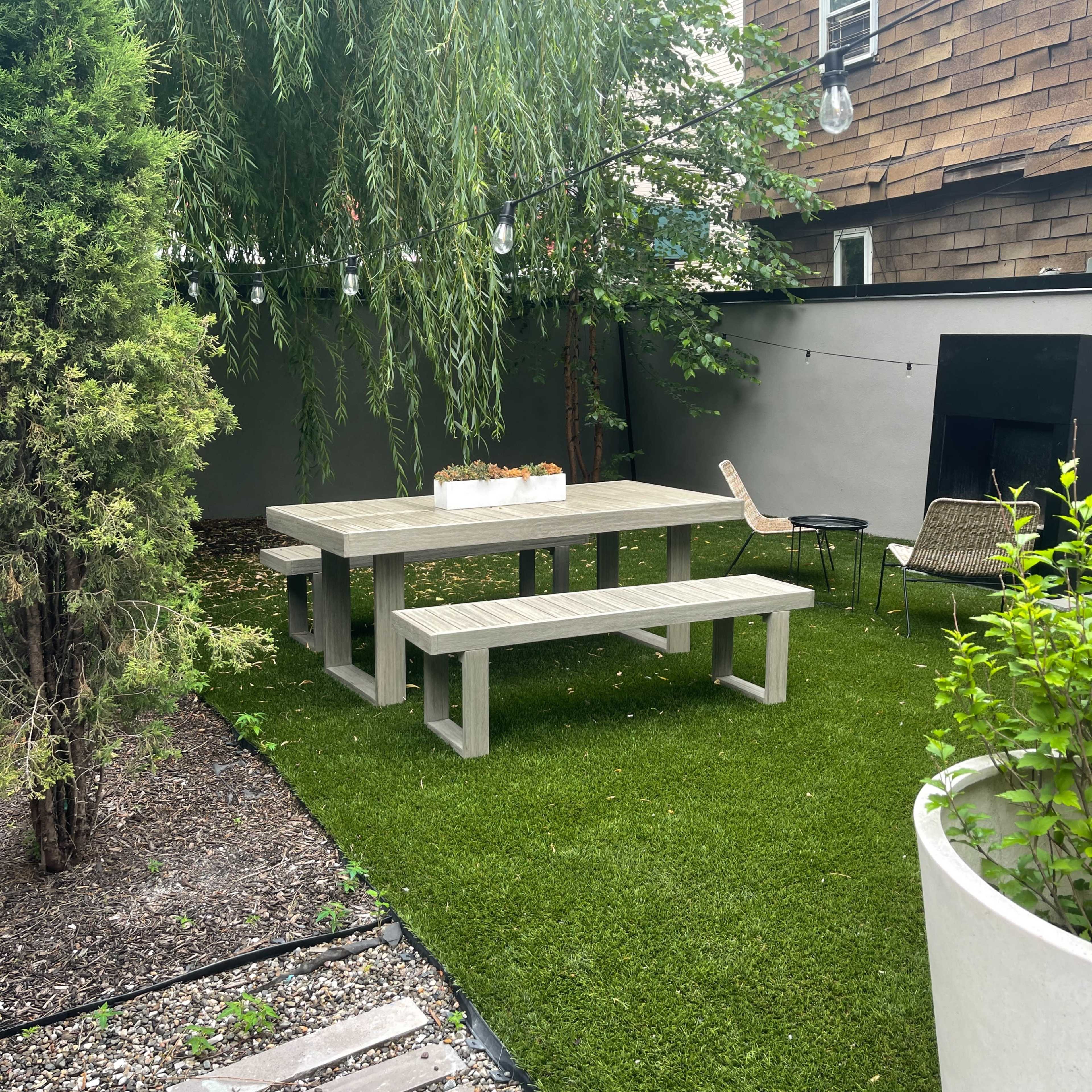 A small outdoor seating area with a wooden picnic table and benches under a weeping willow tree, surrounded by artificial grass and low-maintenance plants.