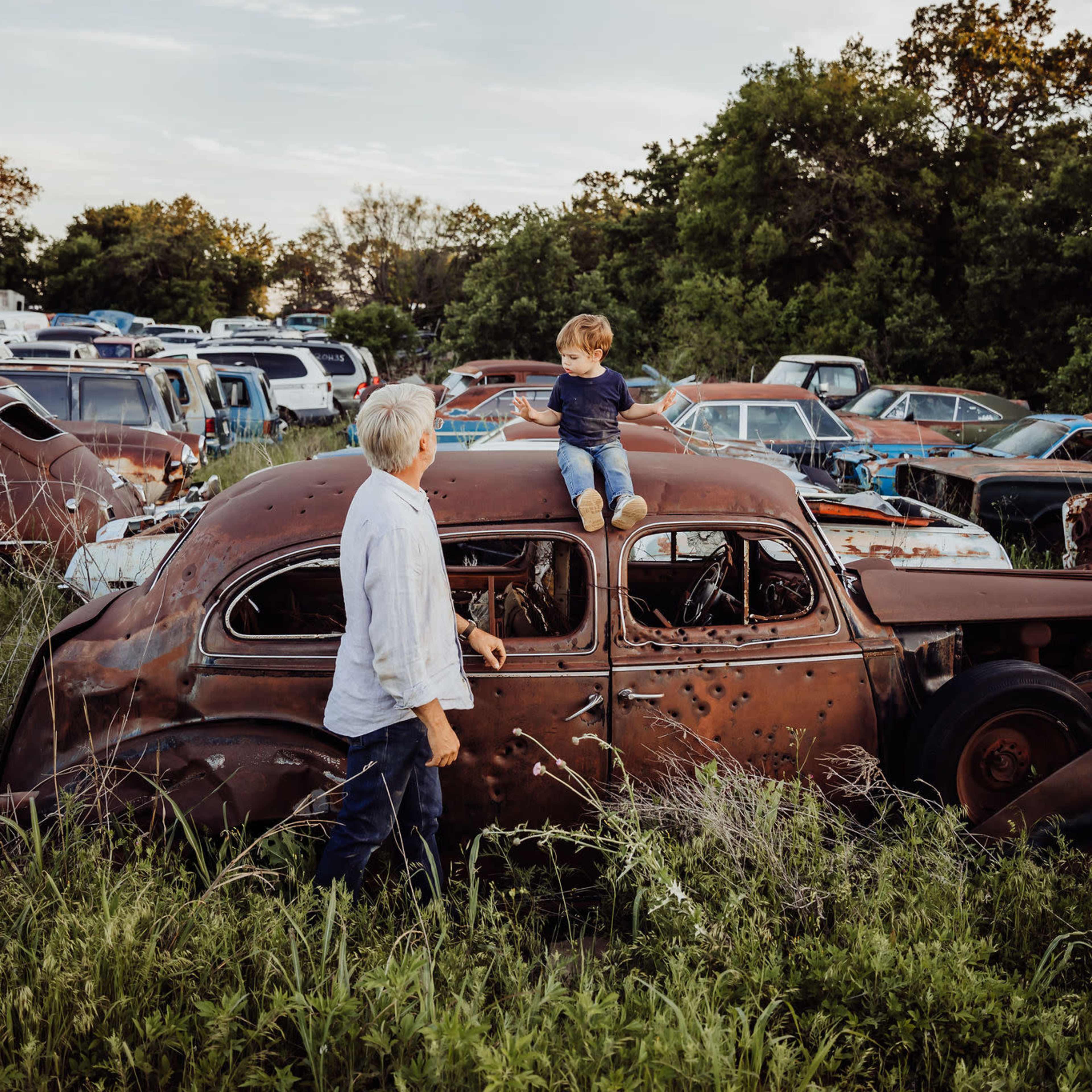 A child sits on the roof of an old, rusted car while an adult stands beside it in a field filled with abandoned vehicles.