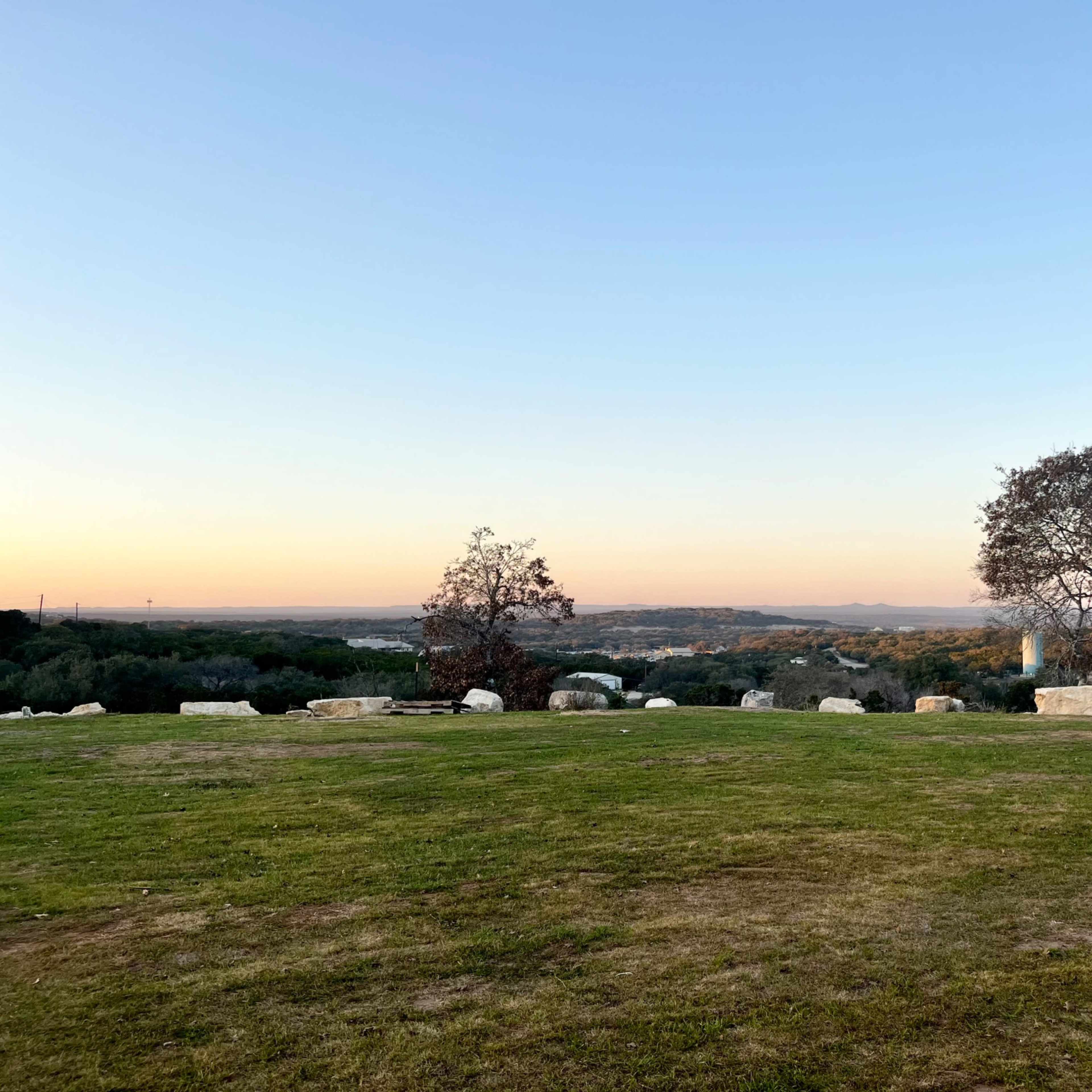 A grassy area with scattered stones, flanked by two trees, overlooking a valley at sunset.