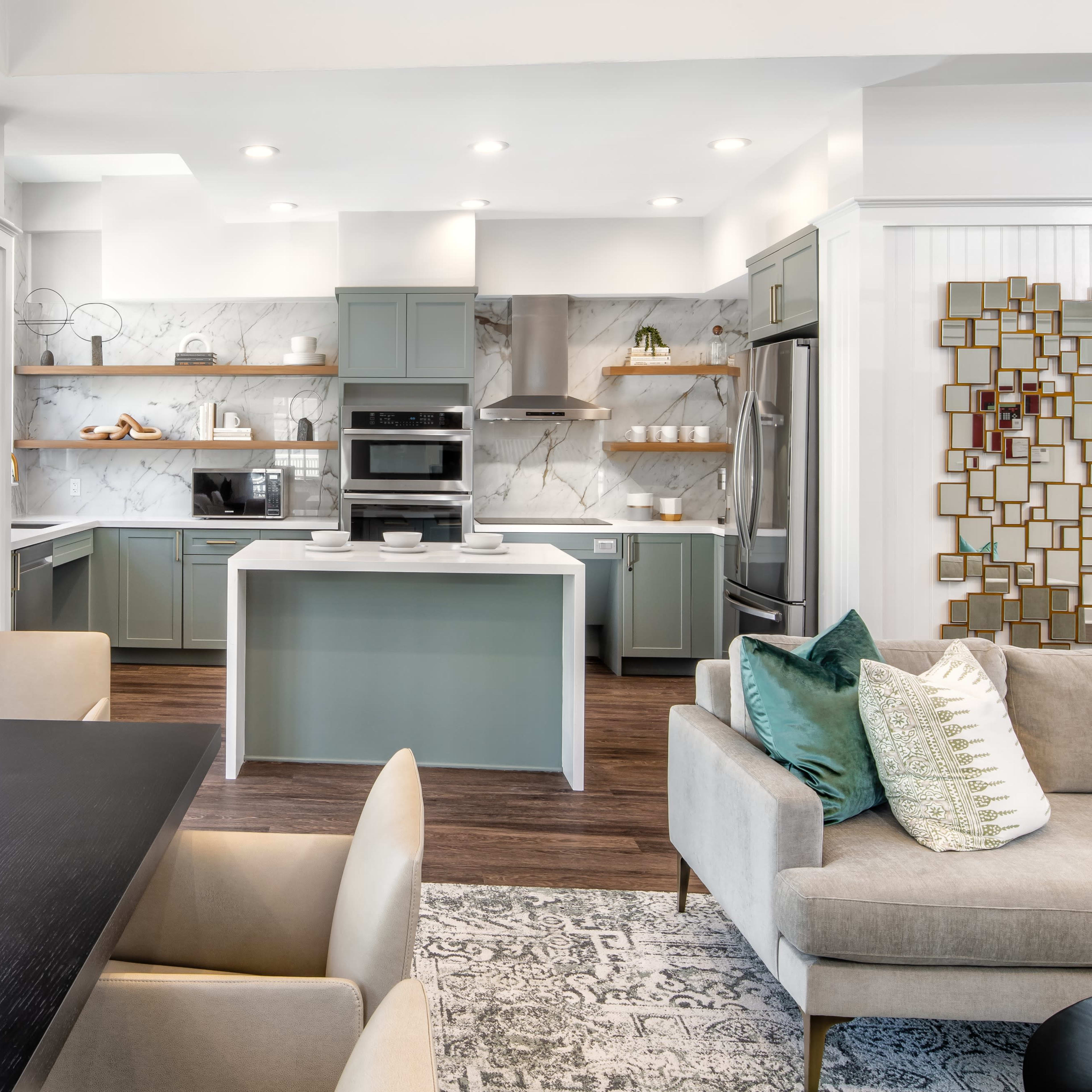 A modern kitchen with light green cabinets, stainless steel appliances, and a dining area featuring a dark wooden table and beige chairs.