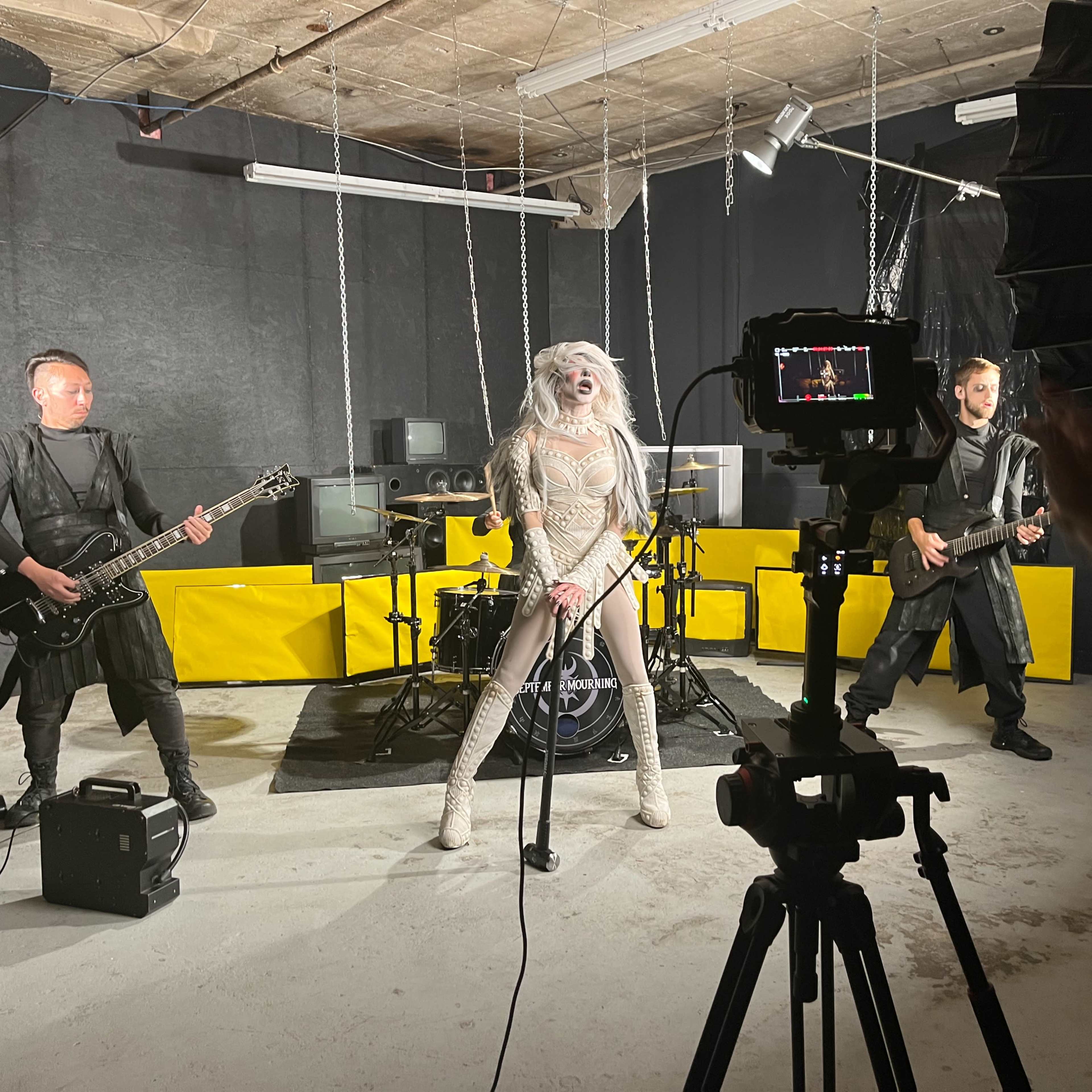 A band performs on stage in a dimly lit room with black walls and yellow accents, with a camera set up to capture the performance.