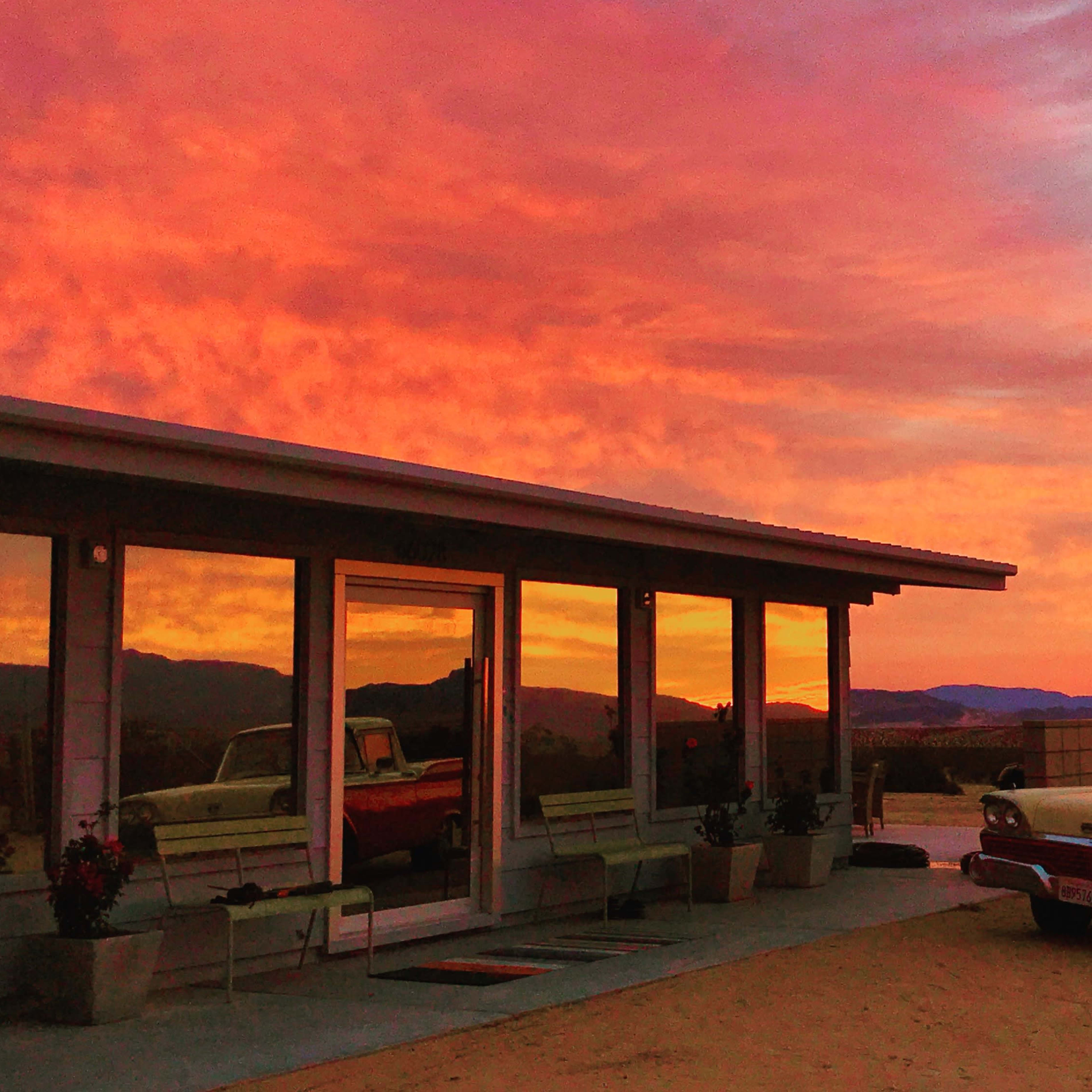 A vintage car is parked in front of a modern building at dusk, with vibrant colors painting the sky behind it.