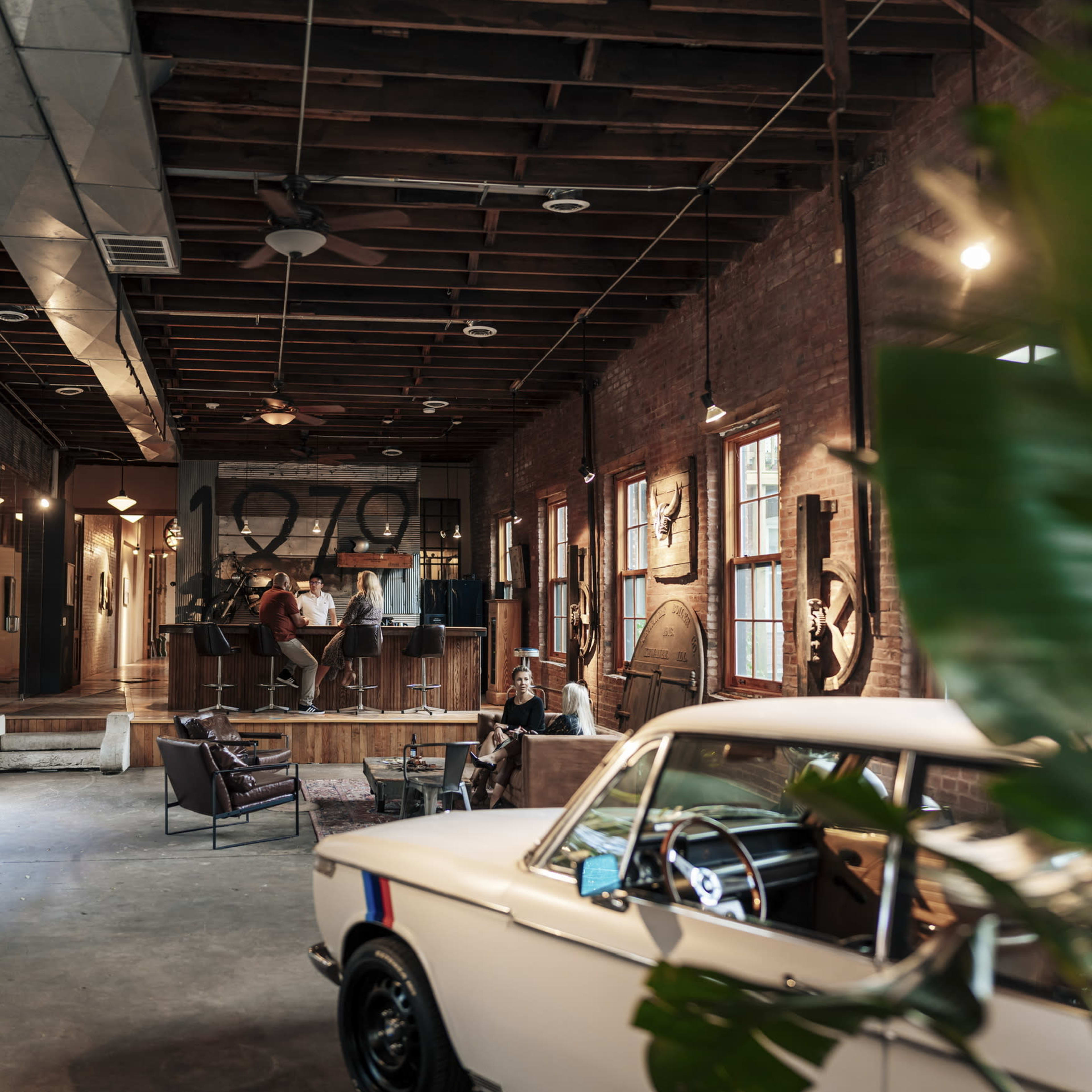 A stylish, industrial-style interior of a café or lounge featuring exposed brick walls, wooden beams, and a classic car on display.