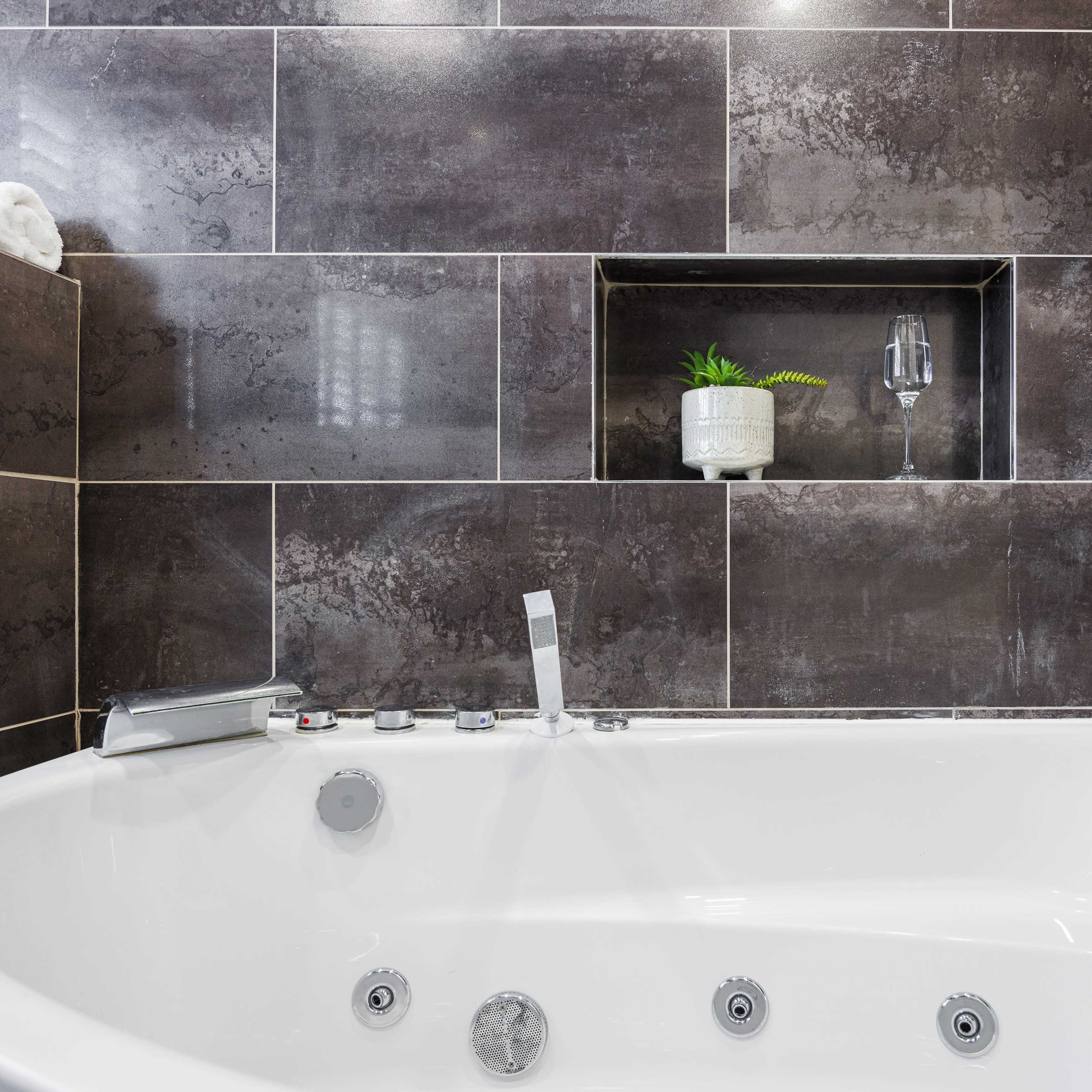 A modern bathroom with a jetted bathtub surrounded by dark tiled walls, including a niche holding a small plant and a glass.
