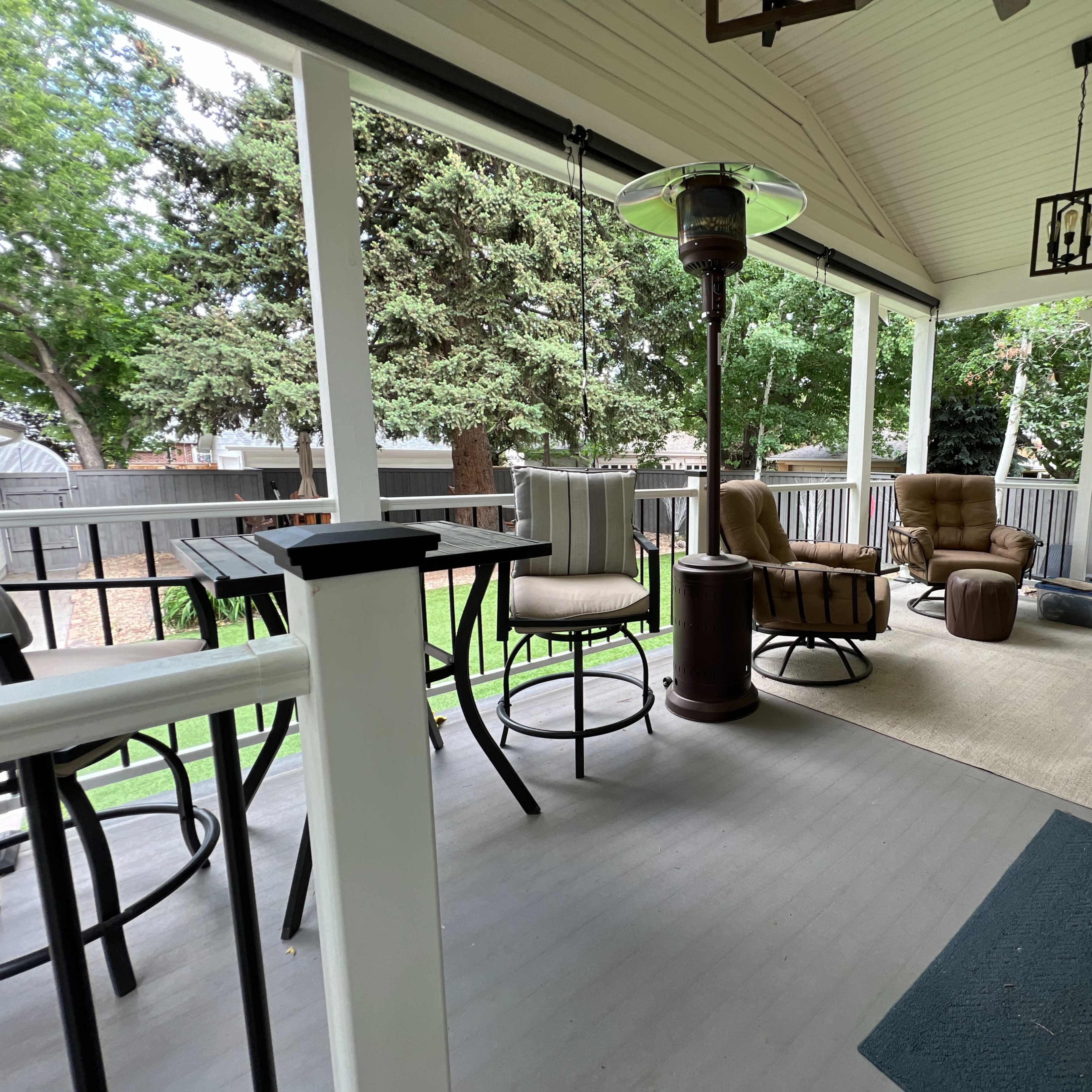 A covered patio featuring various seating options, including a pair of brown reclining chairs, a table with high stools, and a beige rug, surrounded by greenery.