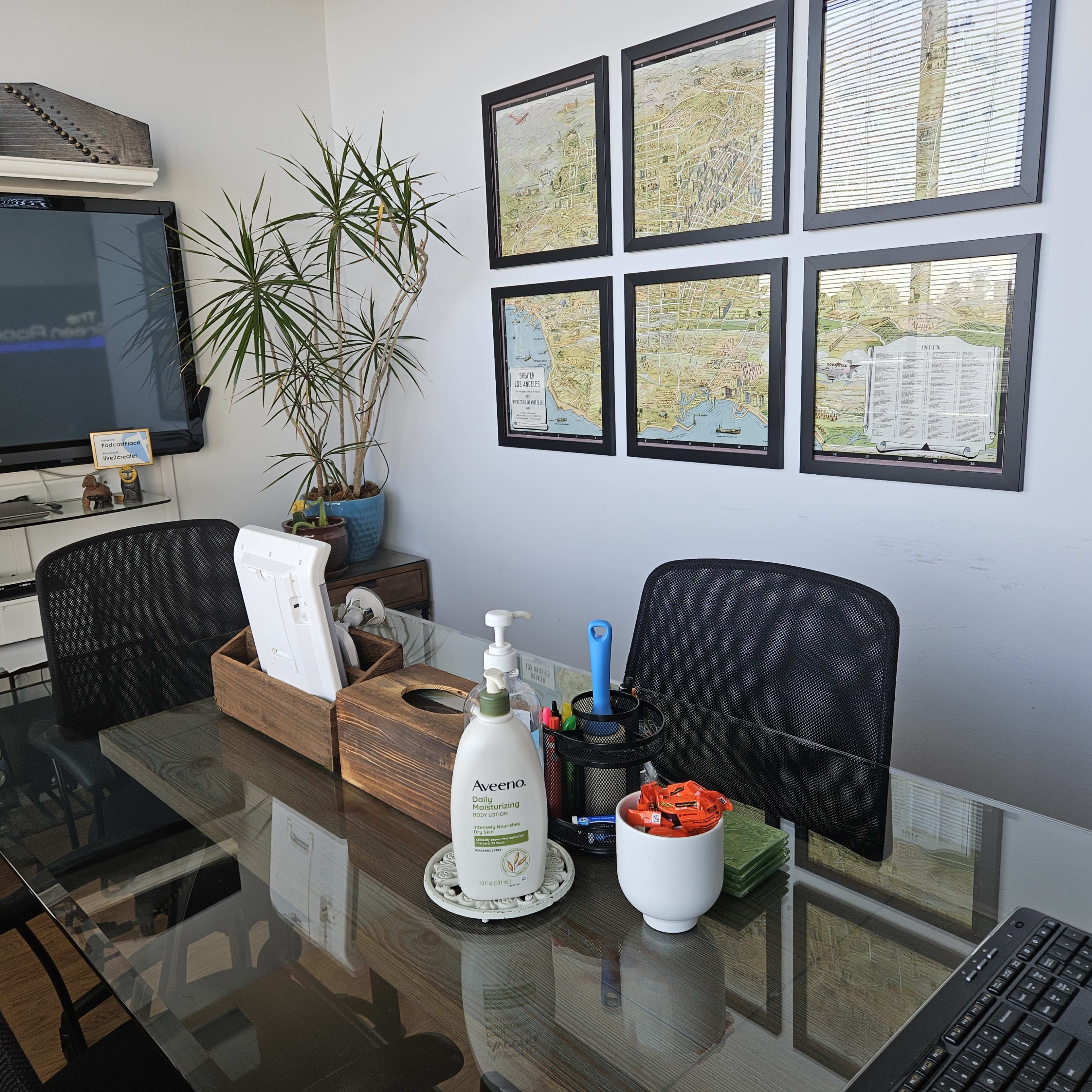 A modern office space featuring a glass desk, a TV on the wall, potted plants, and framed maps.