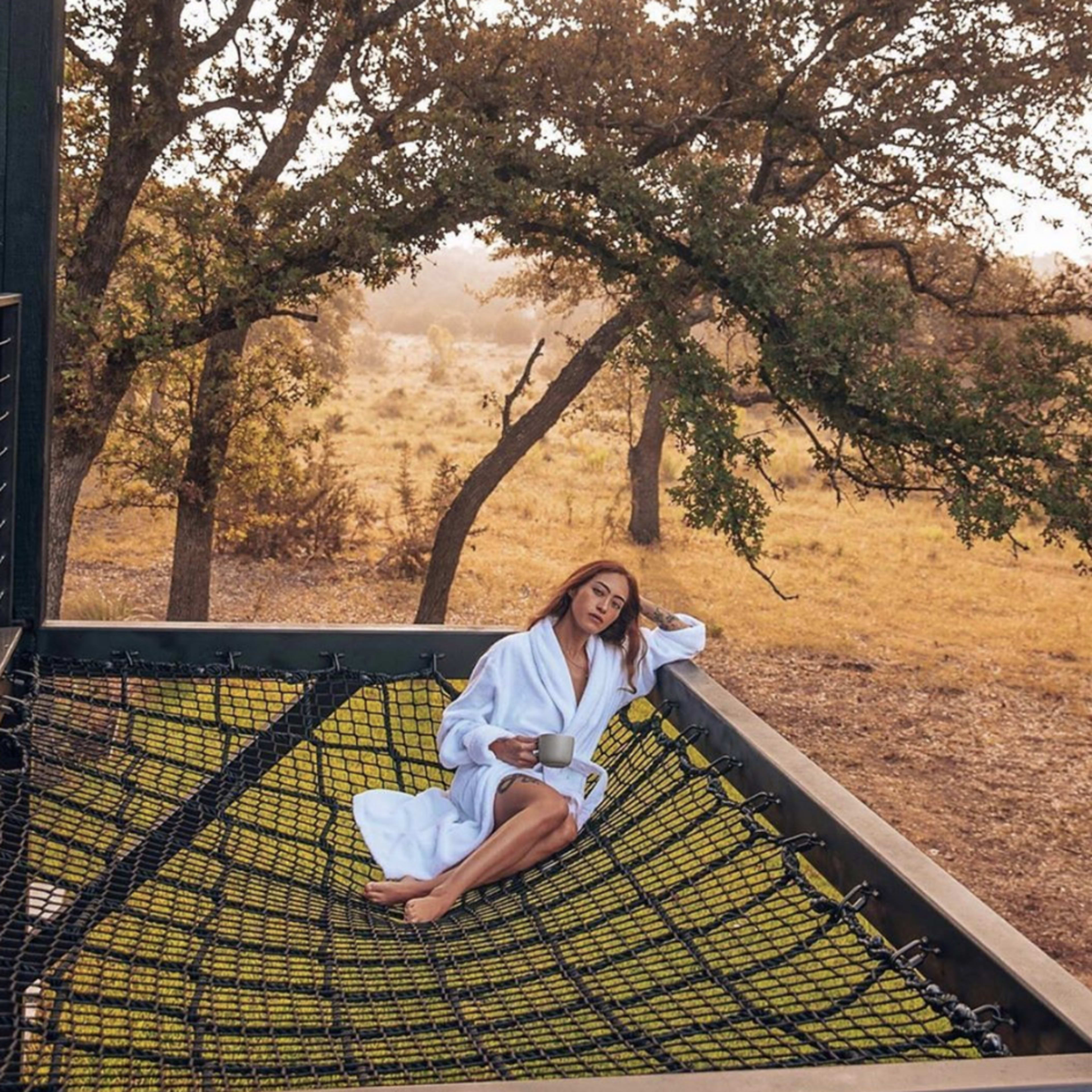 A person in a white robe is sitting on a large net suspended between trees in a natural setting, holding a drink.