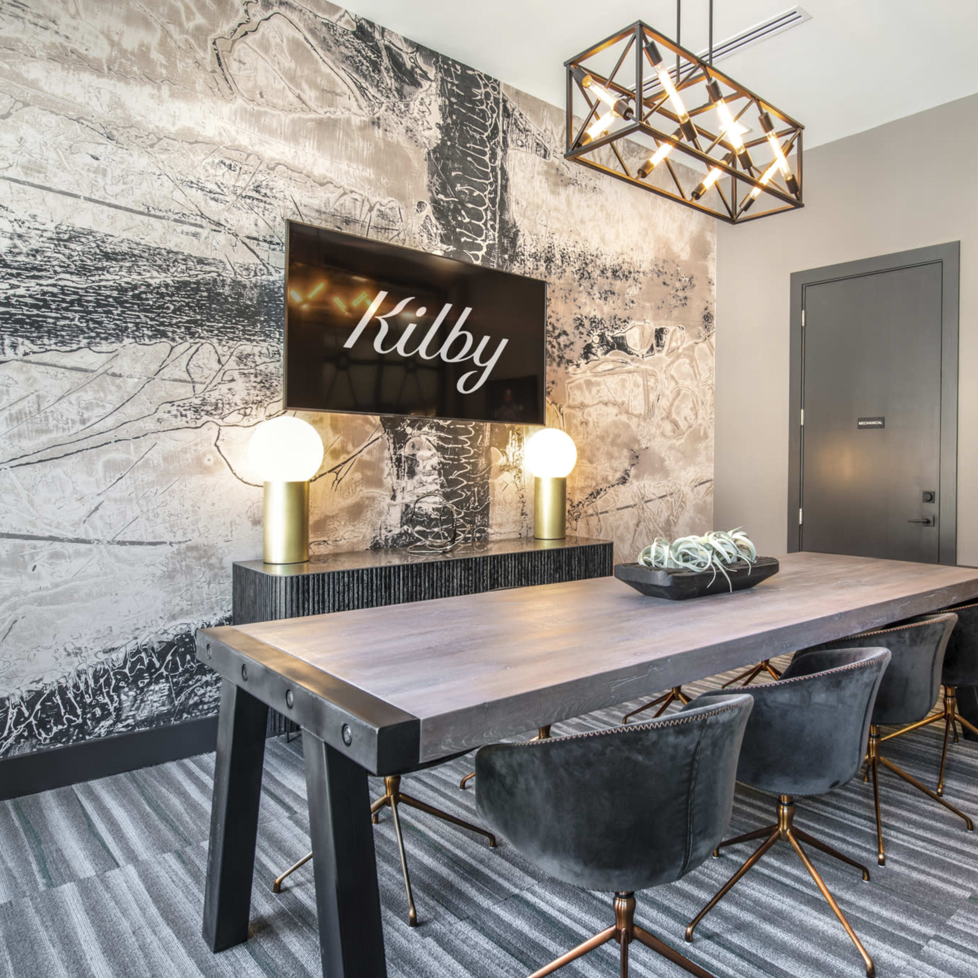 A modern conference room features a large wooden table surrounded by dark chairs, with a decorative wall and a screen displaying the name "Kilby."