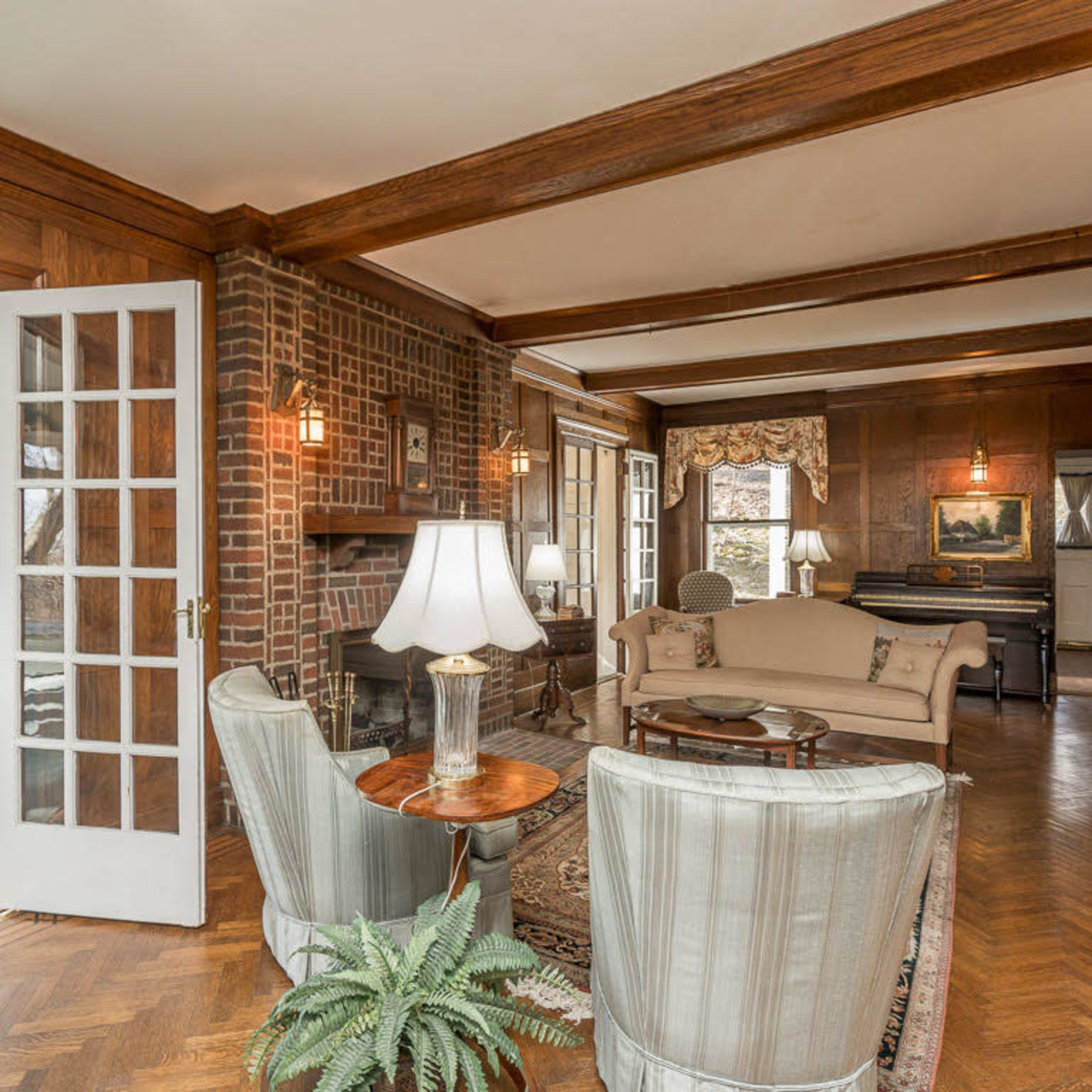 A living room with wooden beams, a brick fireplace, and comfortable seating along with a view into a sunlit room beyond.