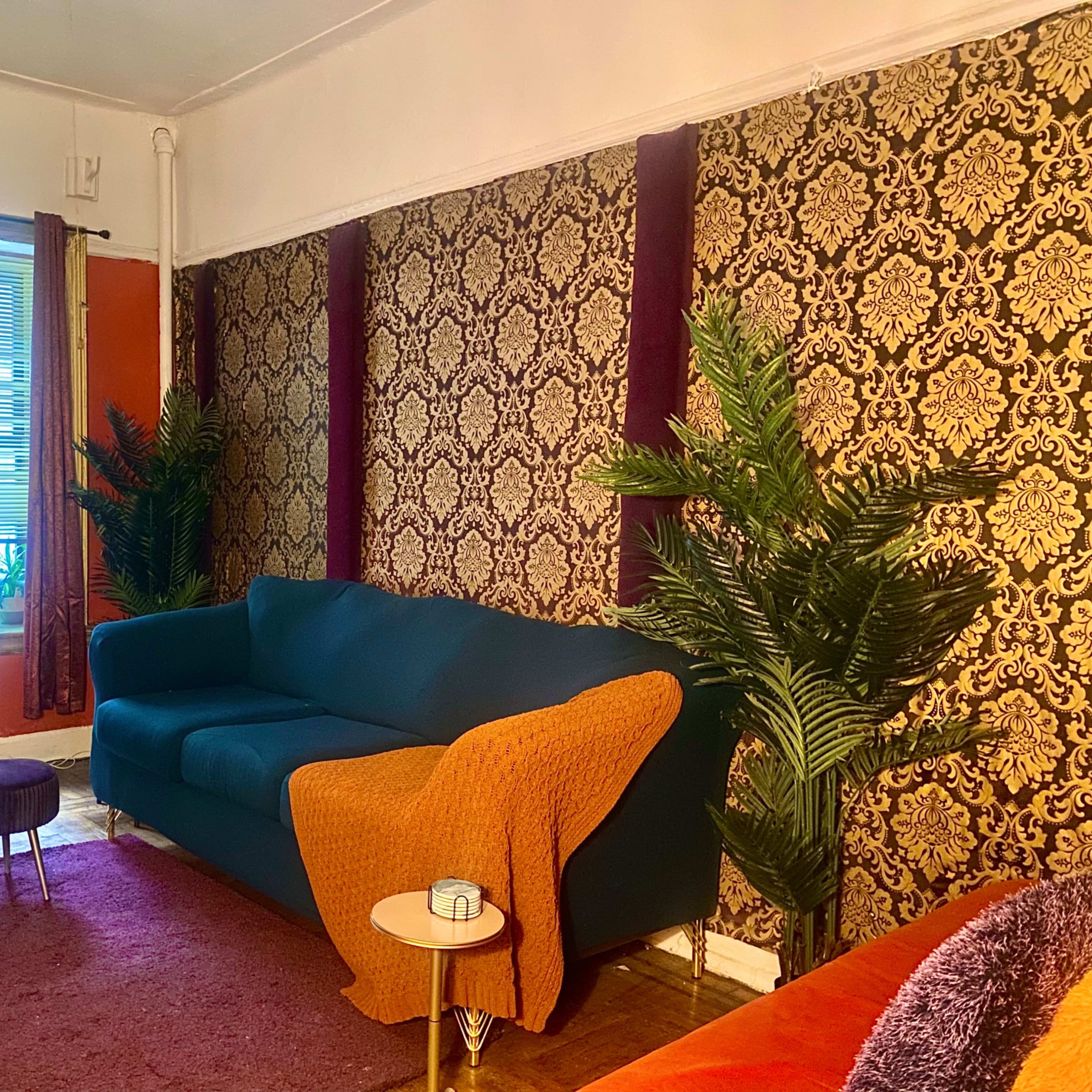 A living room with a blue sofa, a round purple ottoman, patterned wallpaper, and decorative plants.