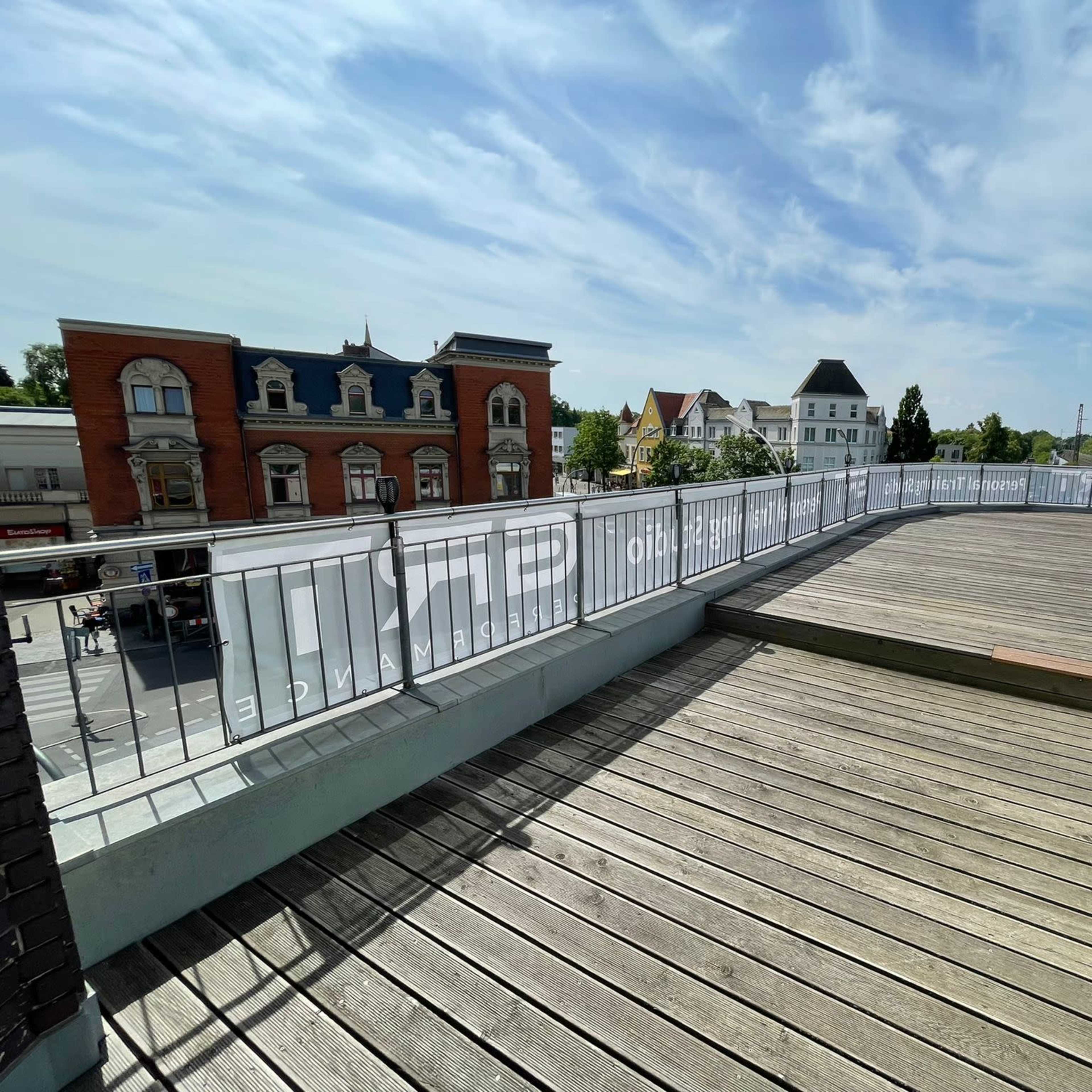 A wooden deck terrace with a railing, overlooking a street lined with buildings and under a partly cloudy sky.