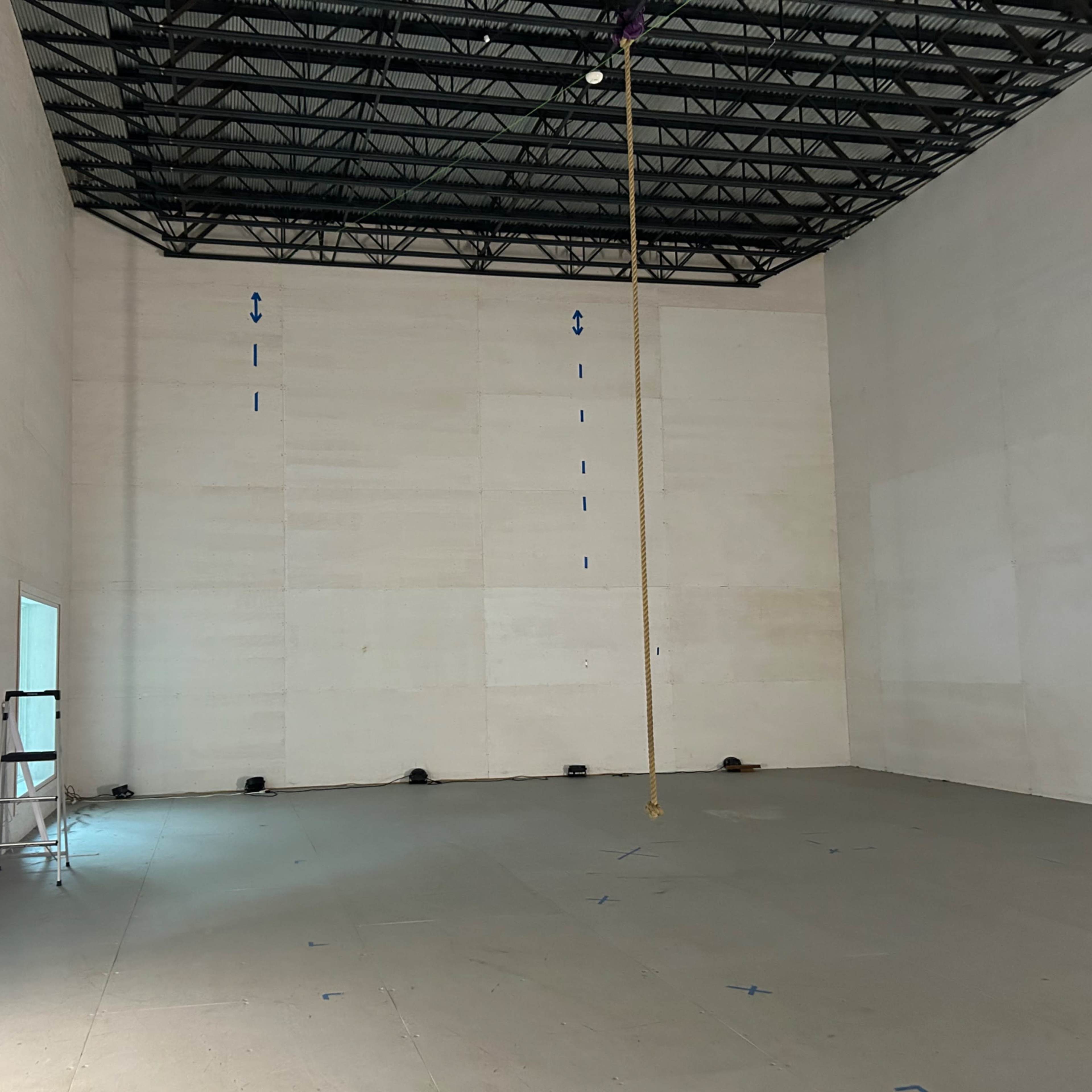 An empty, high-ceilinged interior space with bare walls and a tall rope hanging from the ceiling.