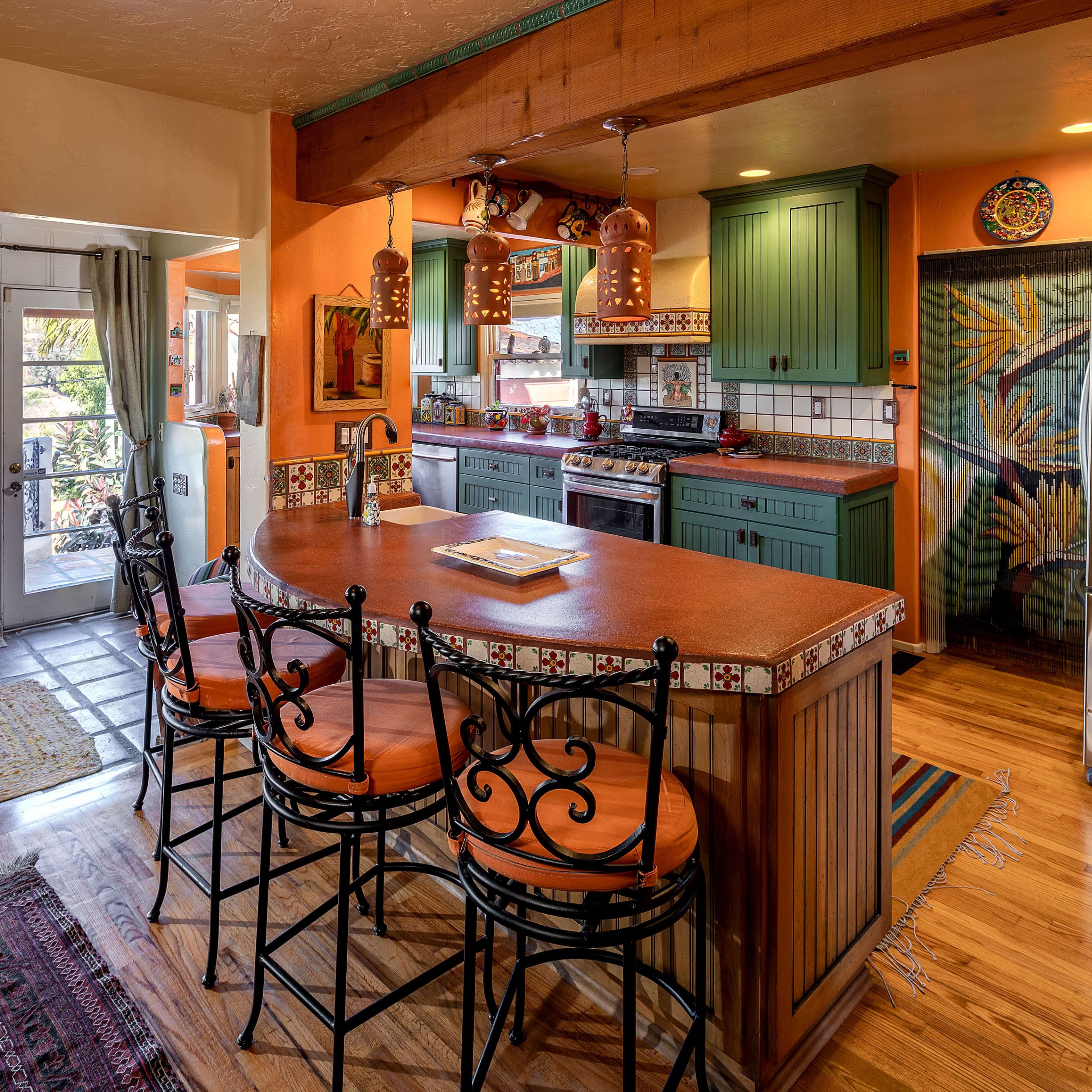A colorful kitchen with wooden cabinetry, a tiled countertop, and a breakfast bar with high-backed chairs, overlooking a living area with a sofa and decorative accents.
