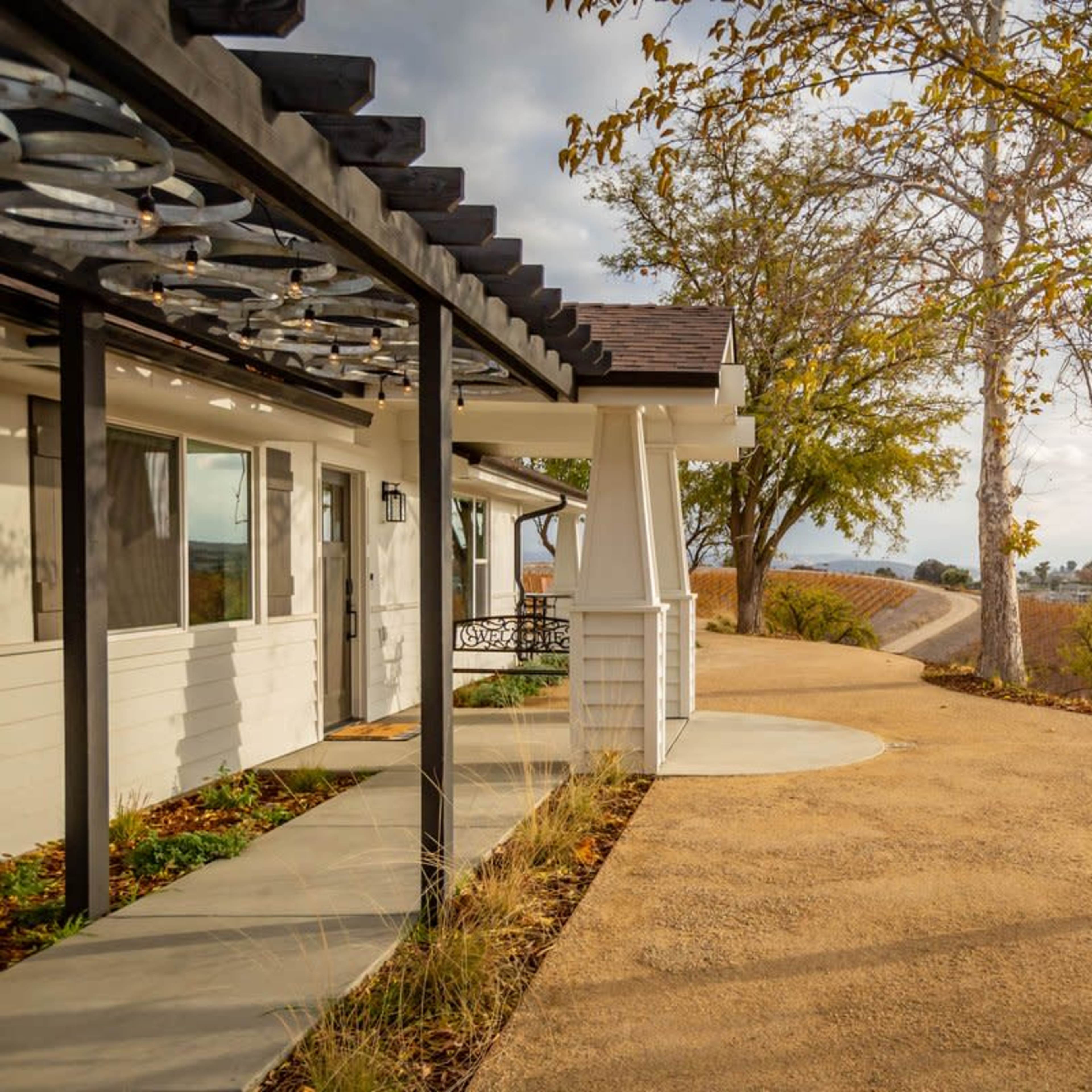 A pathway beside a house lined with trees, leading into a vineyard-covered landscape.