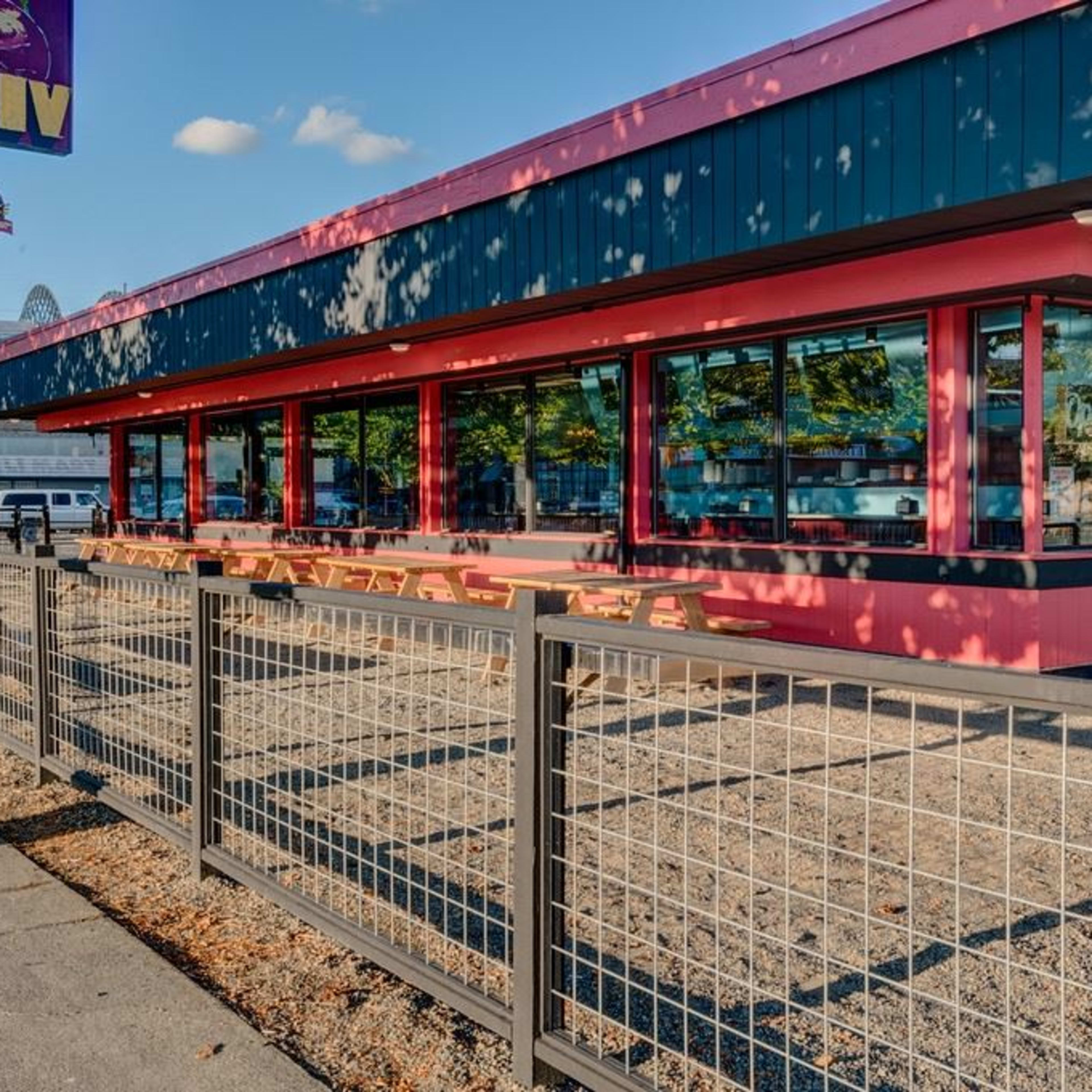 A modern restaurant with large windows and a pink exterior features outdoor seating enclosed by a fence.
