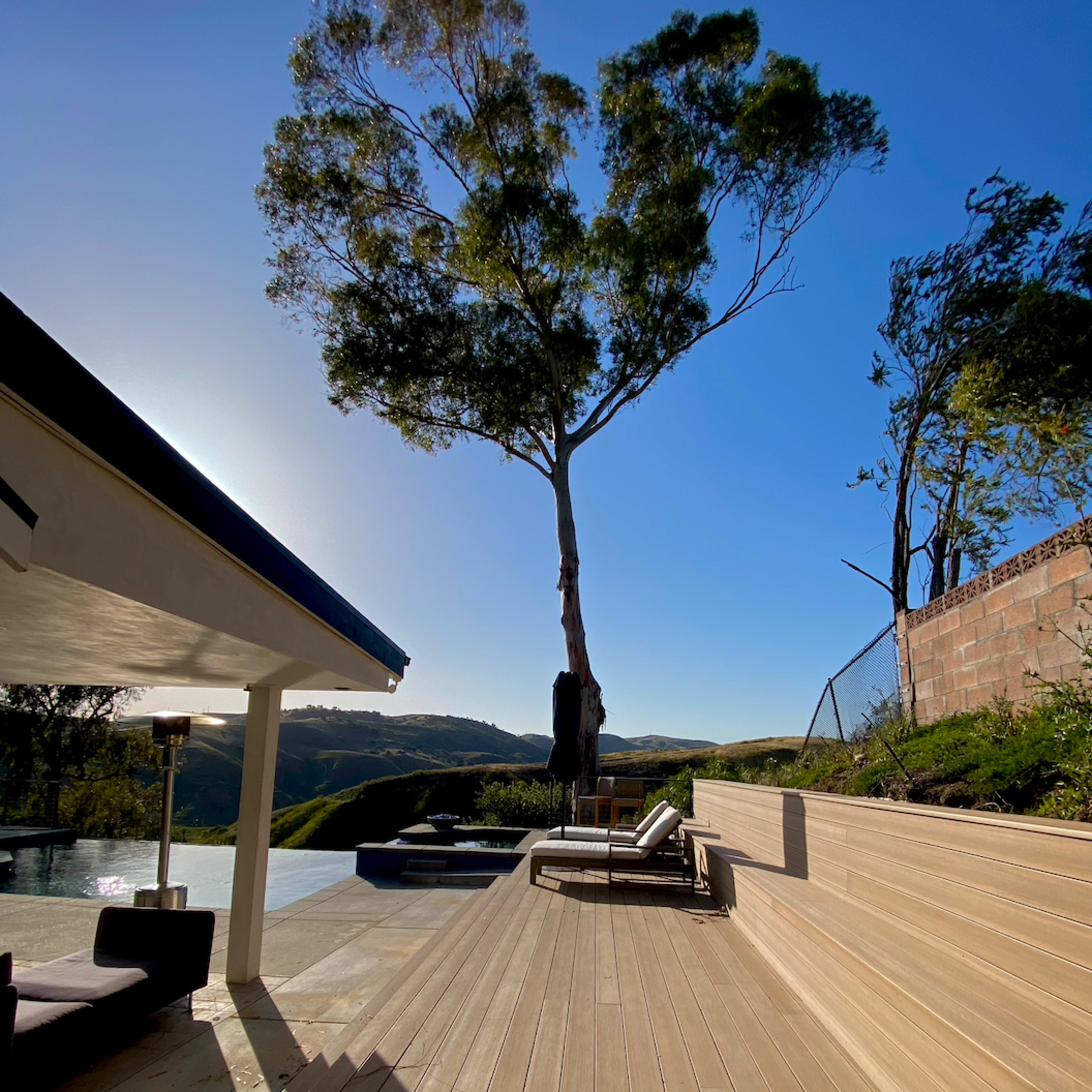 A modern outdoor deck with steps leading down to a pool area, framed by trees and hills under a clear blue sky.