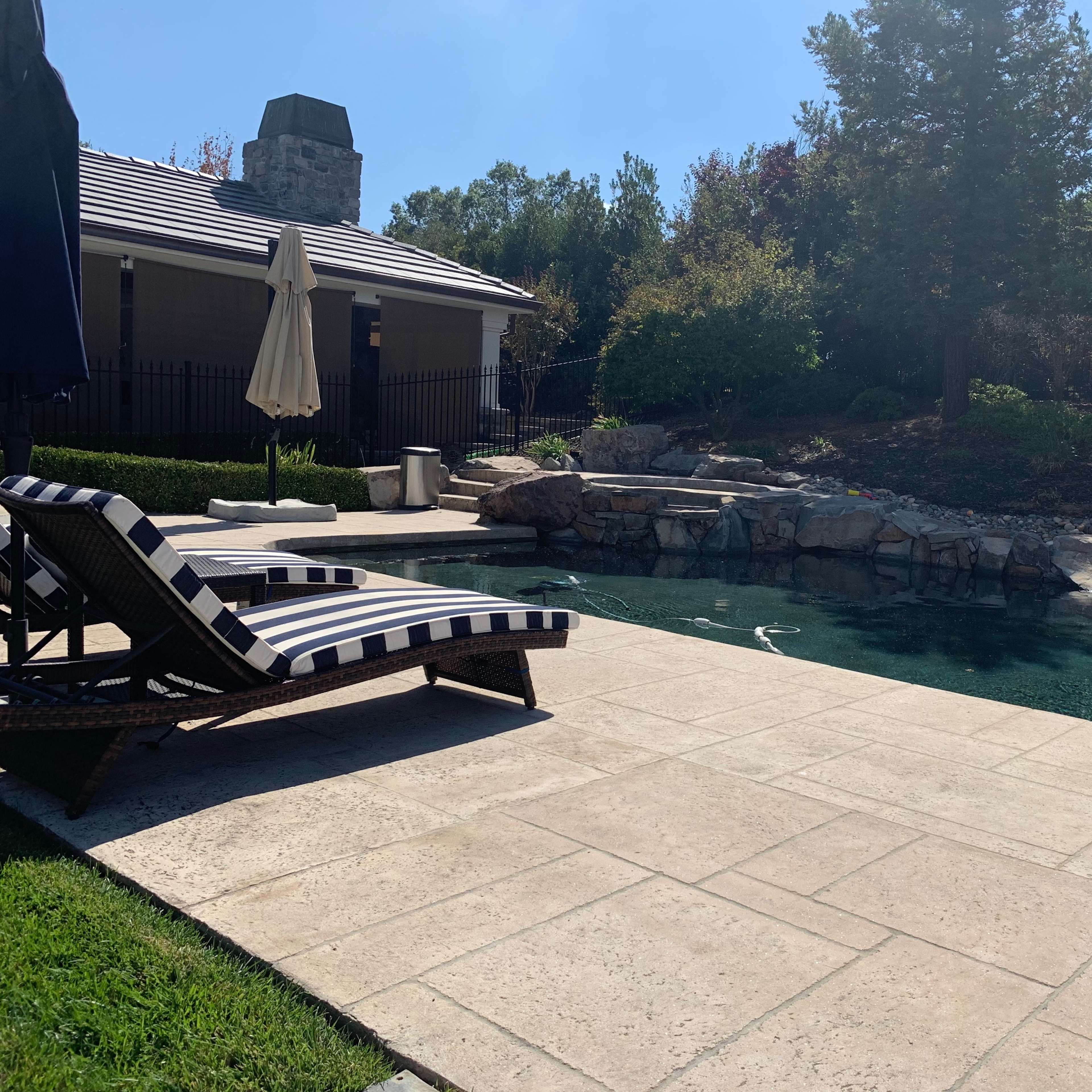 A pool area features a stone patio with a black and white striped lounge chair beside the clear water and landscaped greenery.