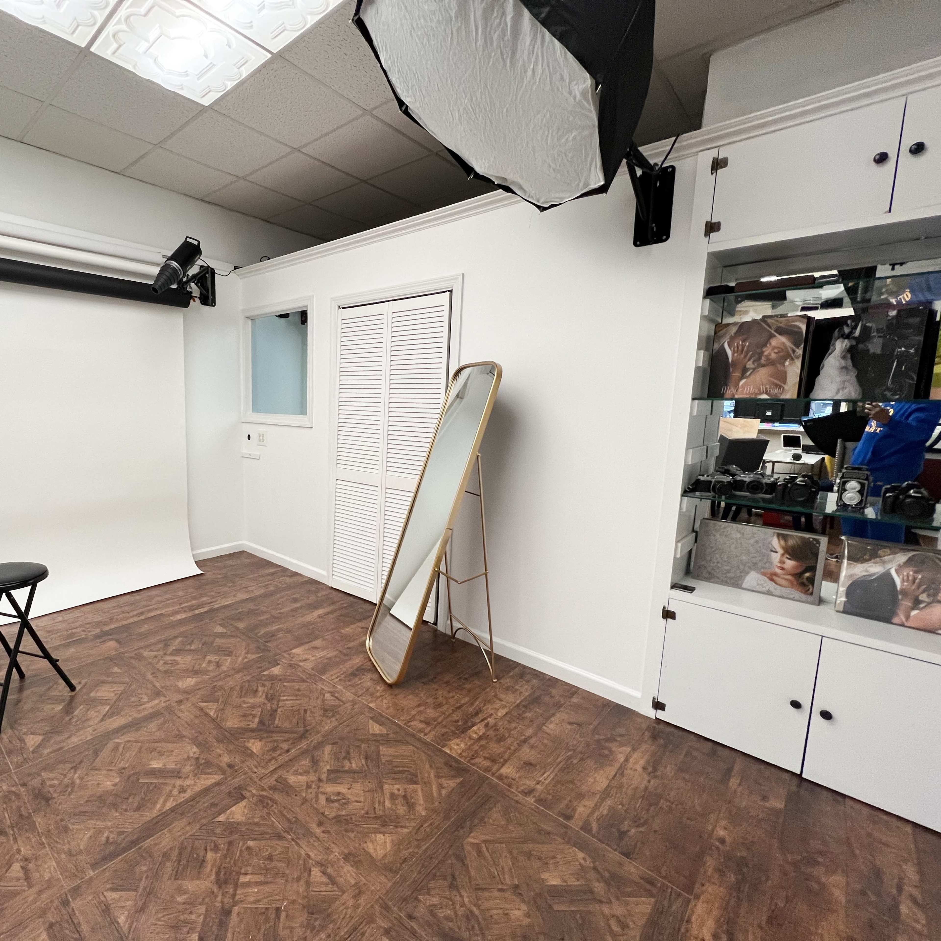 A photography studio with a blank backdrop, a stool in the center, a mirror against the wall, and storage cabinets containing images.