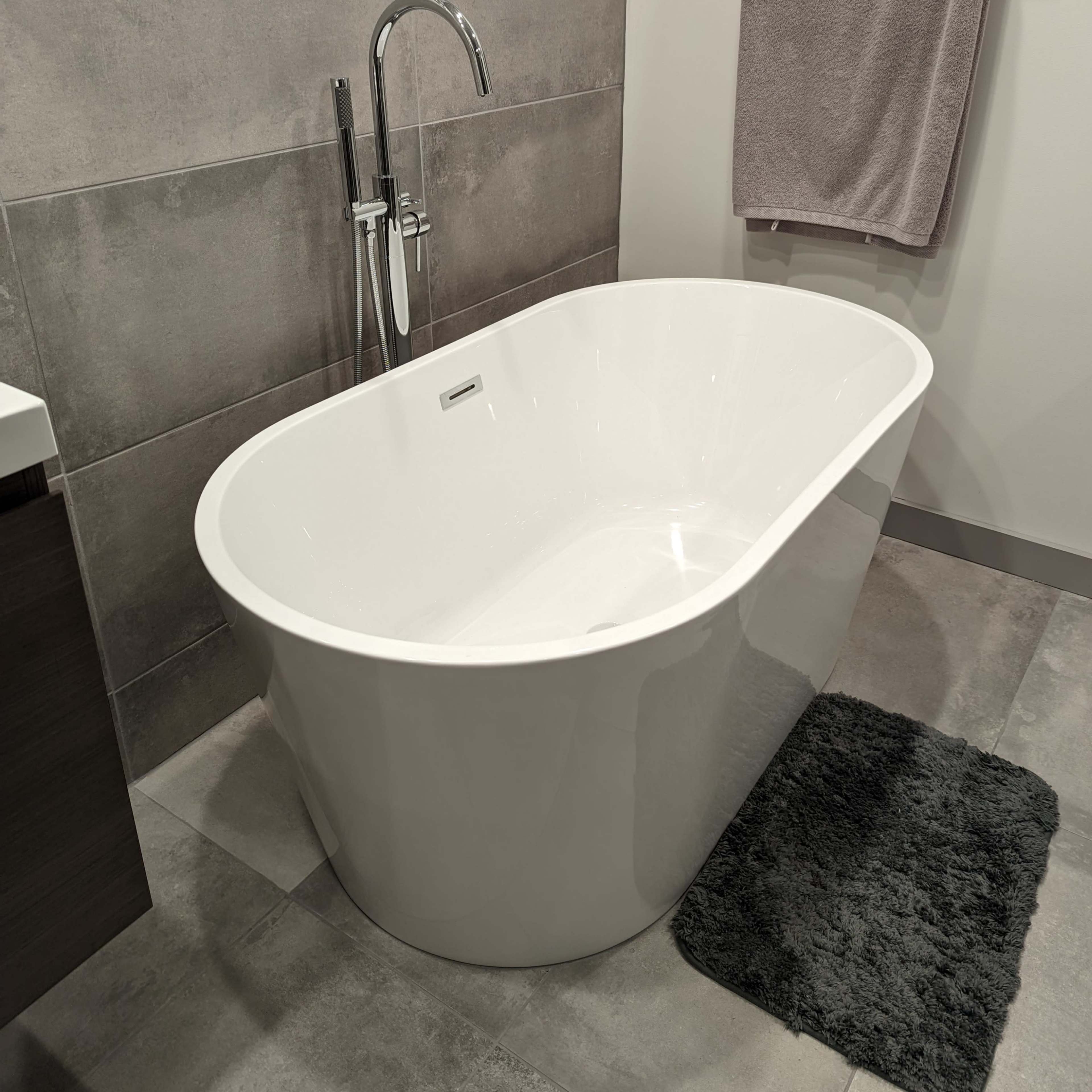 A modern bathroom featuring a freestanding white bathtub next to a towel hanging on a rack and a dark gray rug on the floor.