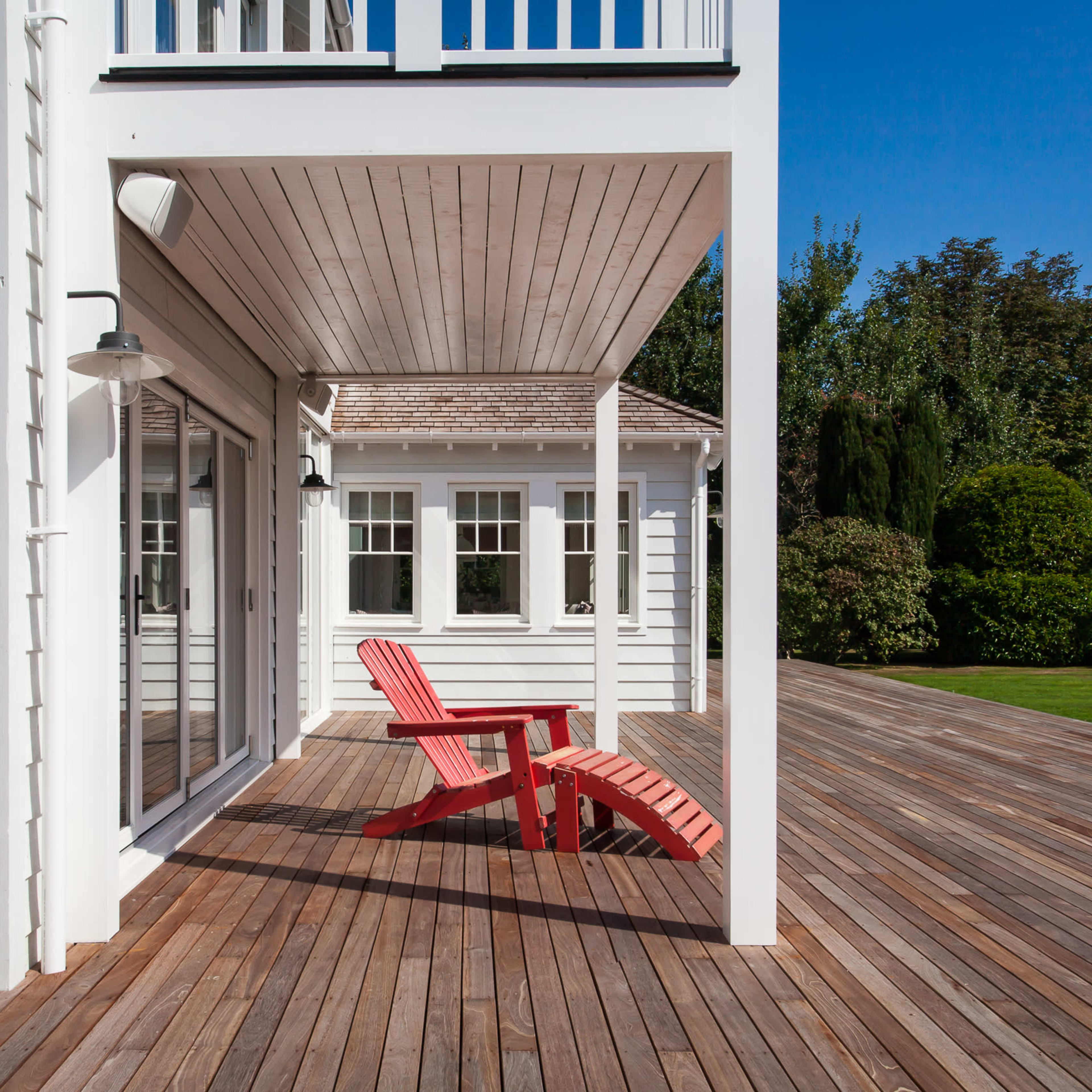 A red adirondack chair sits on a wooden deck next to a light-colored house with large glass doors and windows.