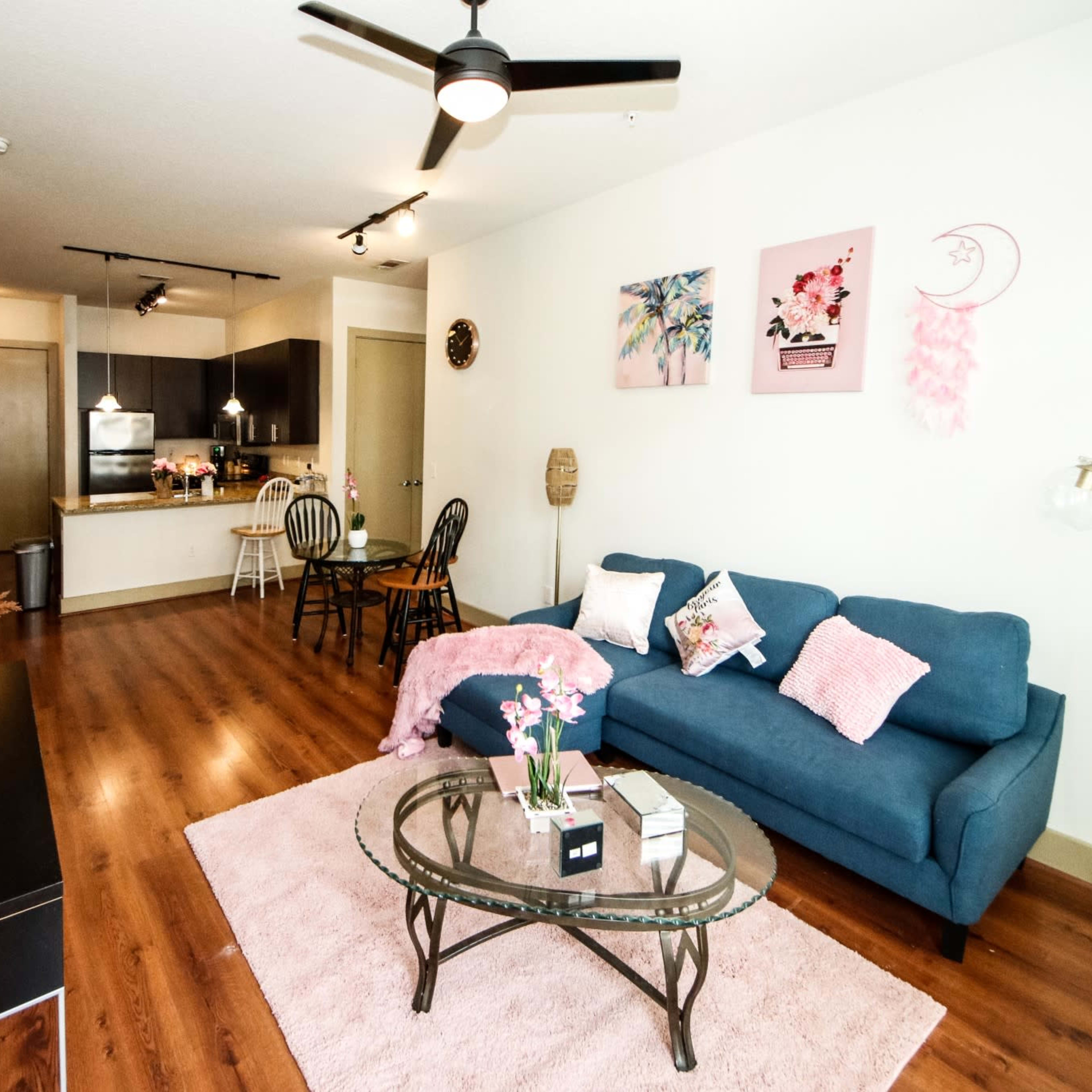 A modern living room with a blue sofa, a glass coffee table, and a kitchen area in the background, adorned with decorative wall art.