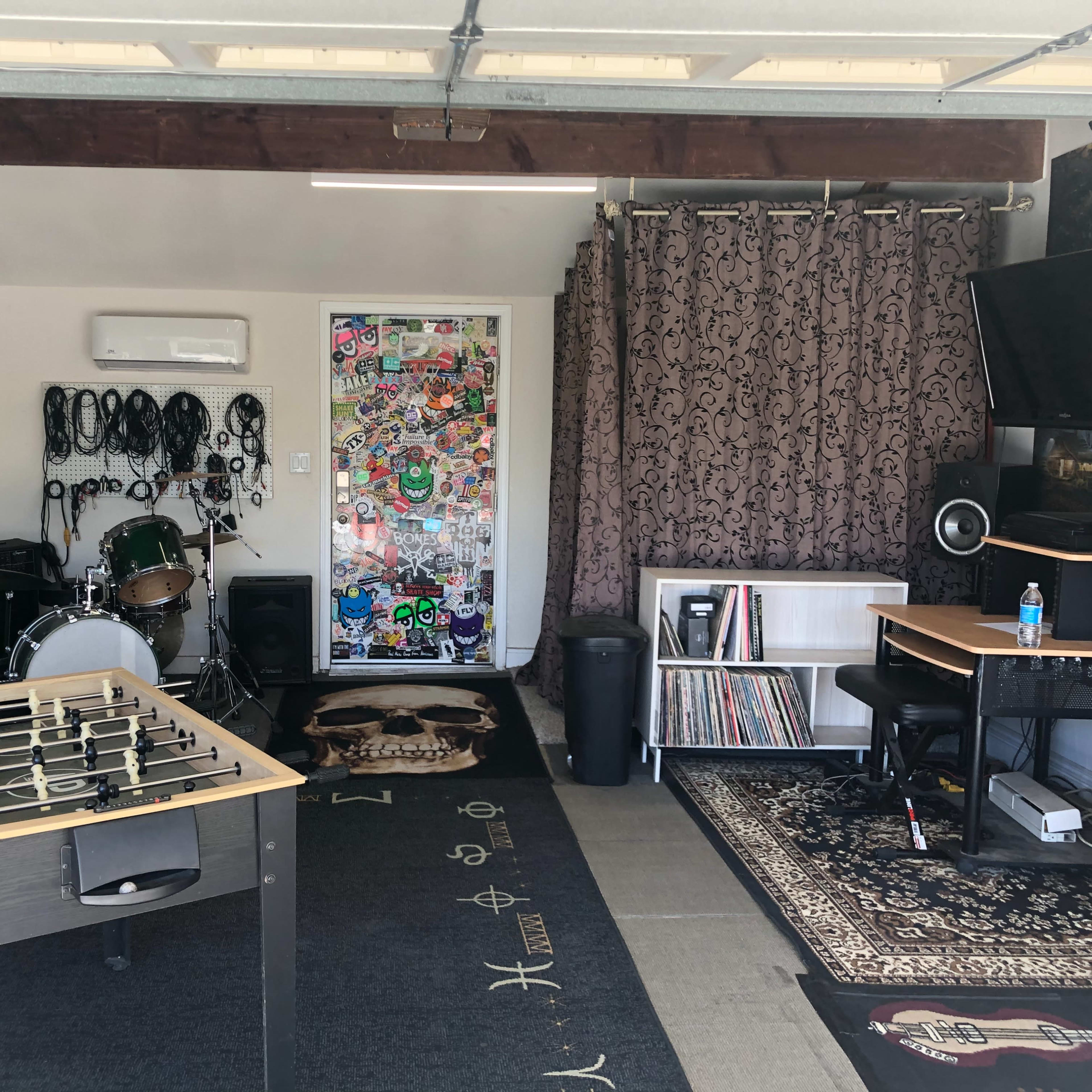 A room with a foosball table, a drum set, a desk with a computer, and a wall adorned with stickers and cables.