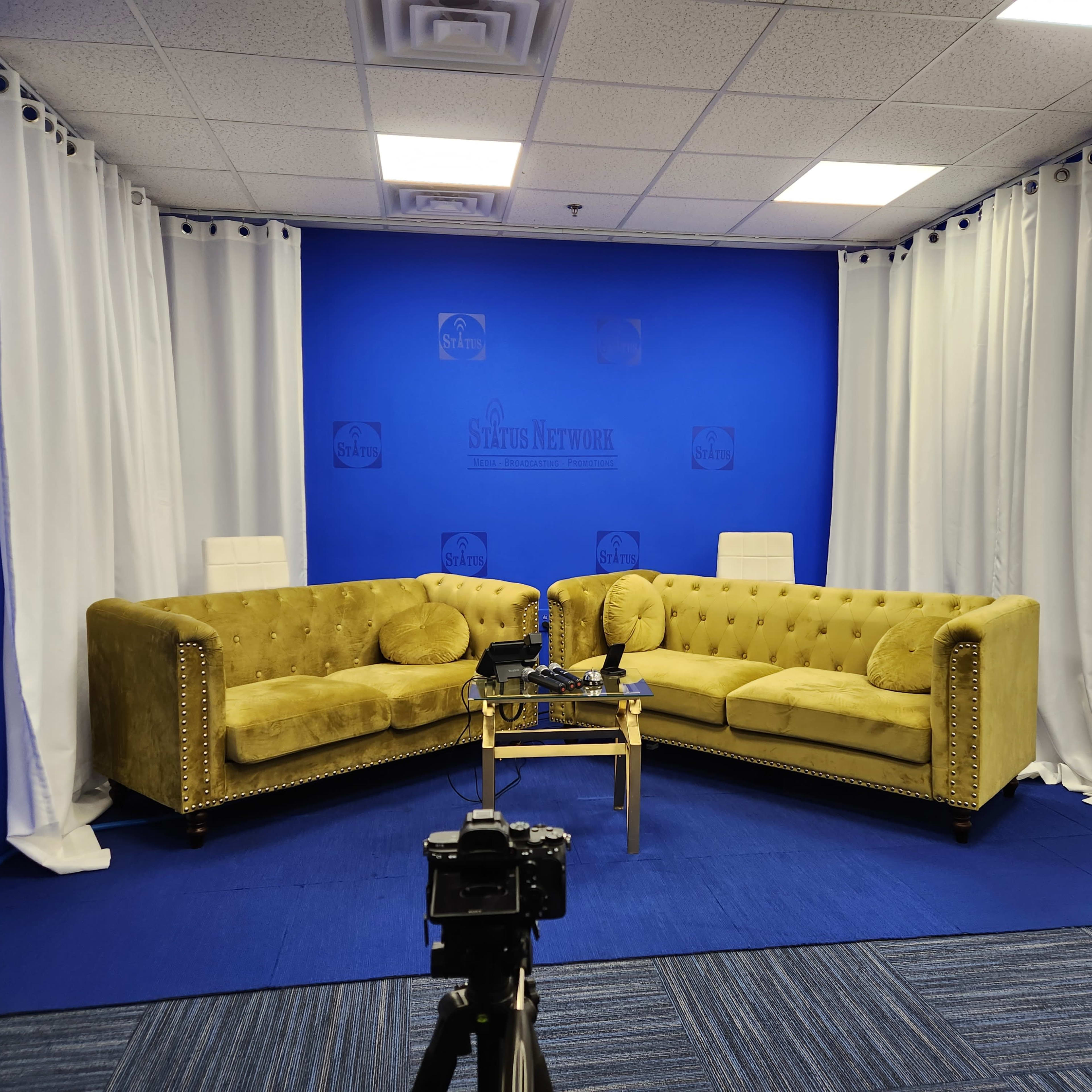 A television studio setup with two yellow tufted sofas facing each other on a blue carpet, surrounded by white curtains and a blue backdrop.