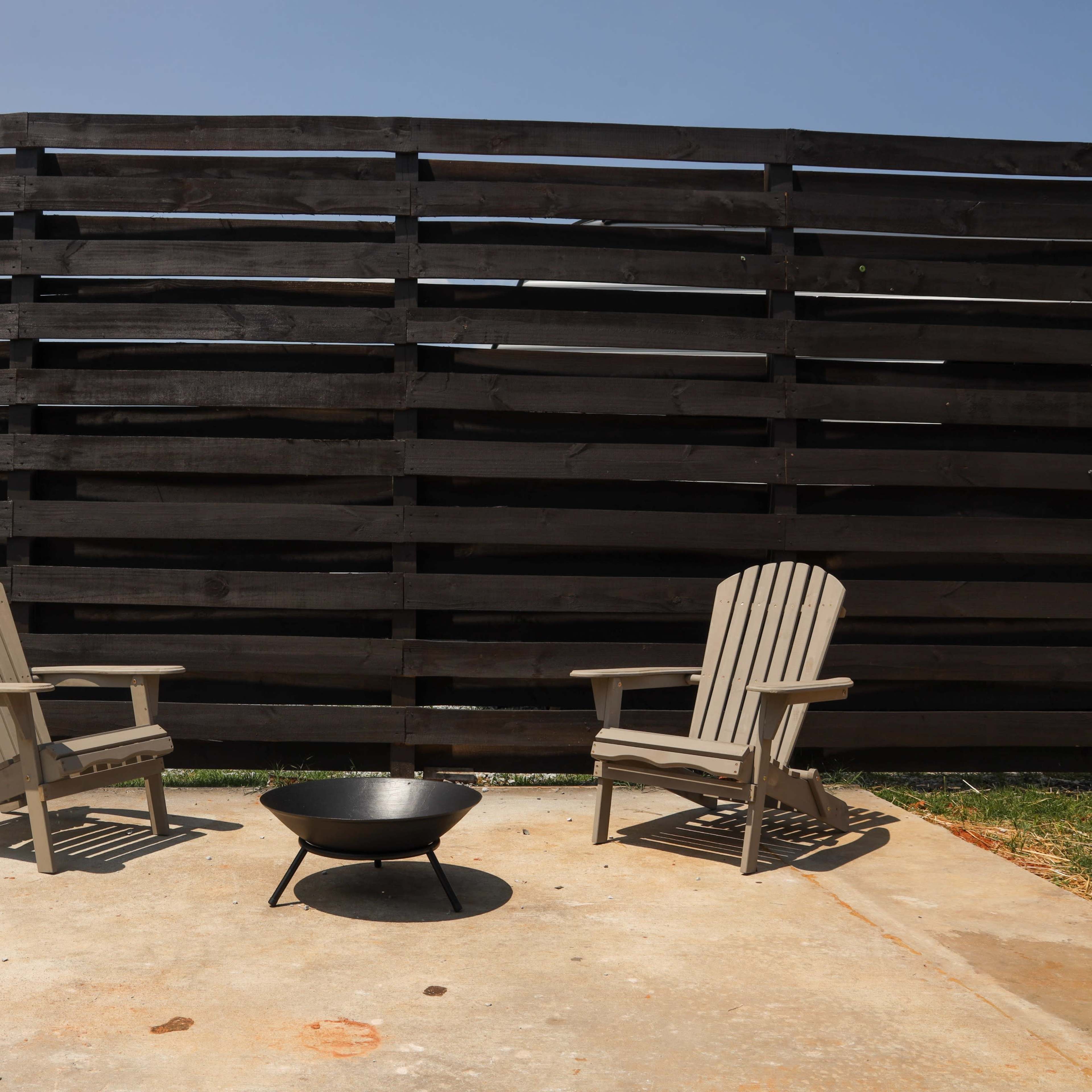 Two wooden chairs are positioned in front of a fire pit on a concrete area next to a tall wooden fence.