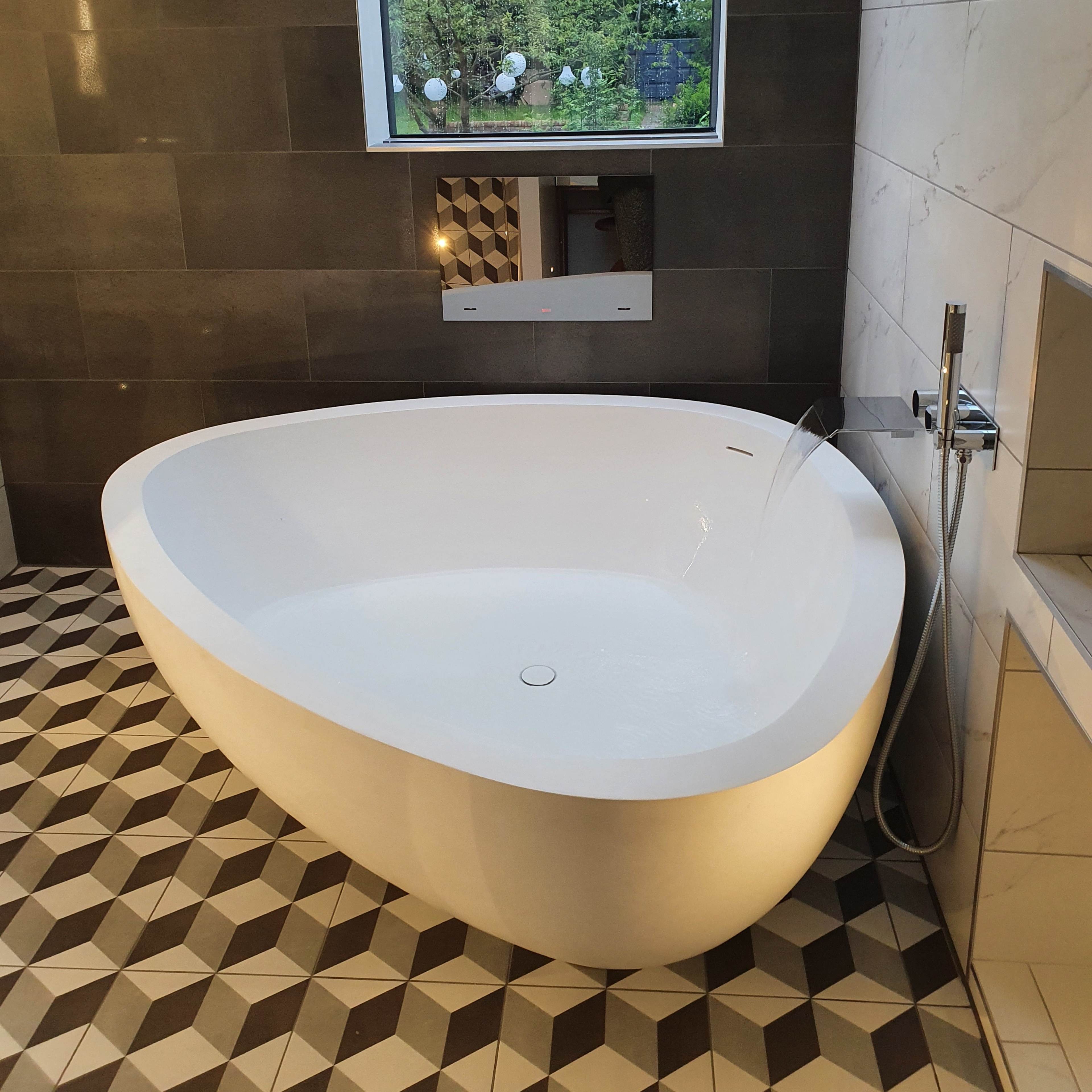 A freestanding oval bathtub is positioned in a bathroom with a large window overlooking greenery, set against a patterned tile floor and gray walls.