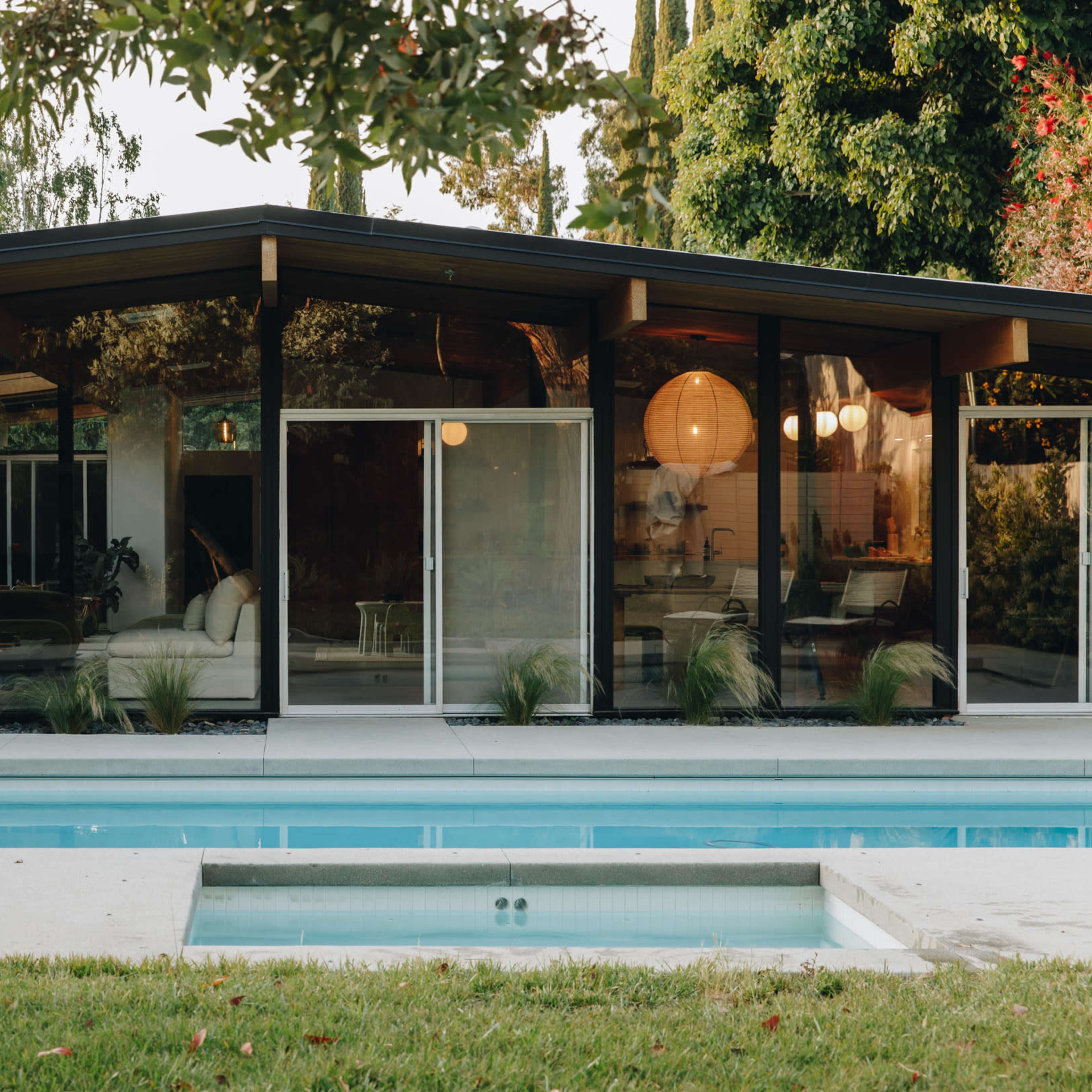 A modern, black house with large glass windows overlooks a swimming pool and is surrounded by greenery.