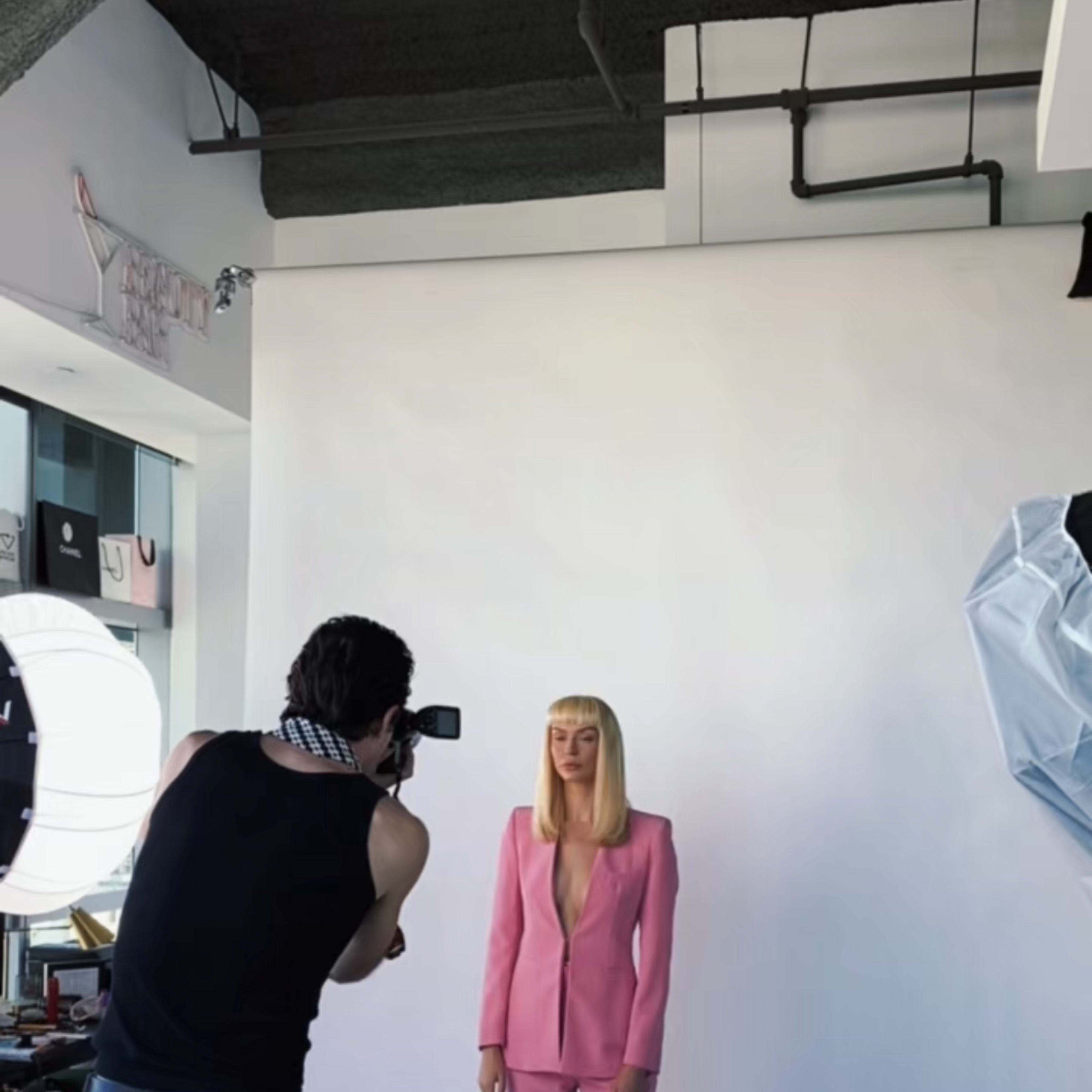 A photographer captures a model dressed in a pink suit against a plain white backdrop in a studio setting.