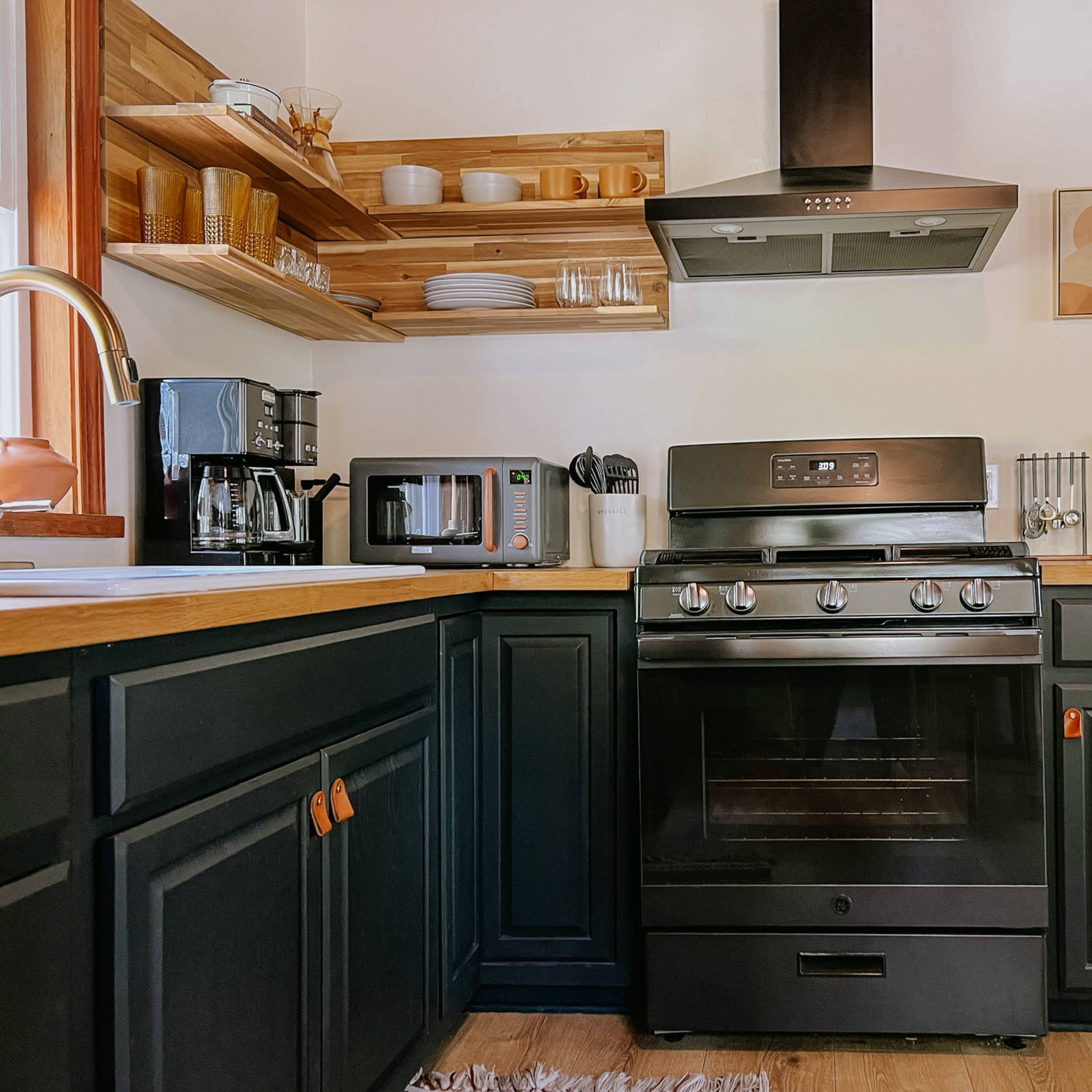 A modern kitchen features dark cabinets, a stainless steel stove, a microwave, and wooden shelves displaying dishes and coffee utensils.