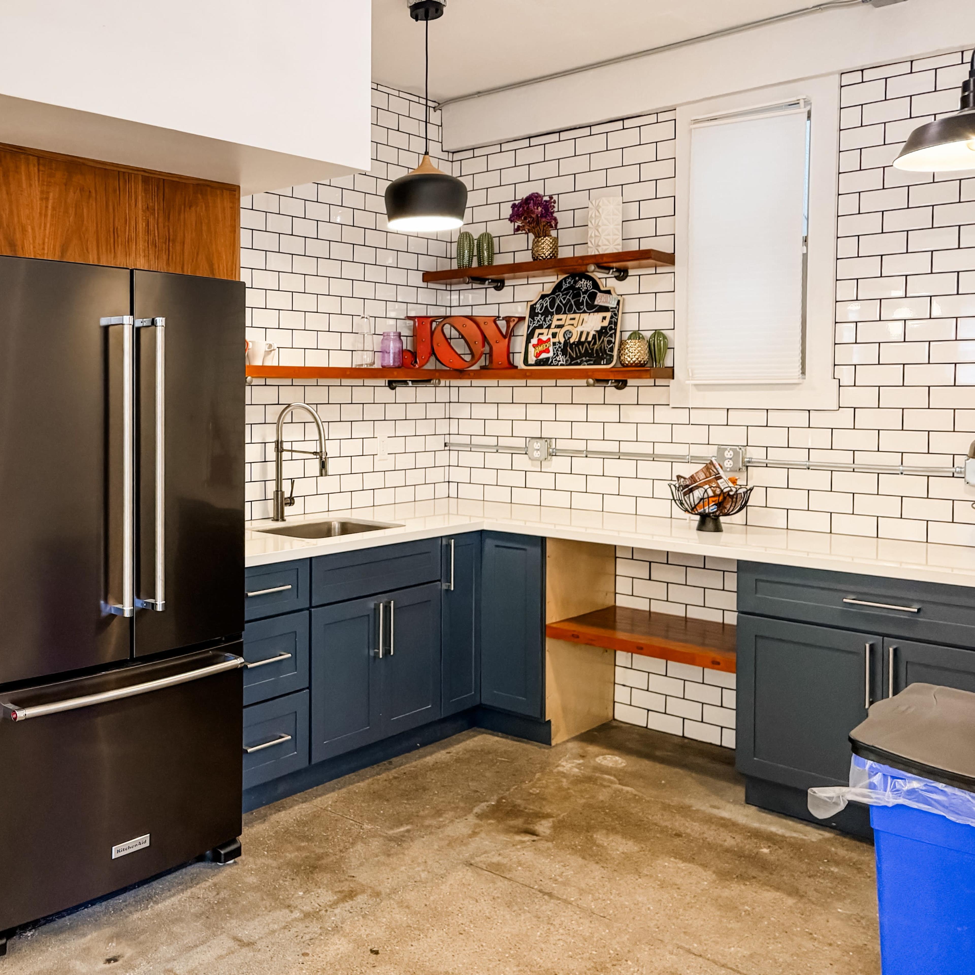 A modern kitchen featuring a black refrigerator, wooden shelving, gray cabinets, a white countertop, and subway tile backsplash.