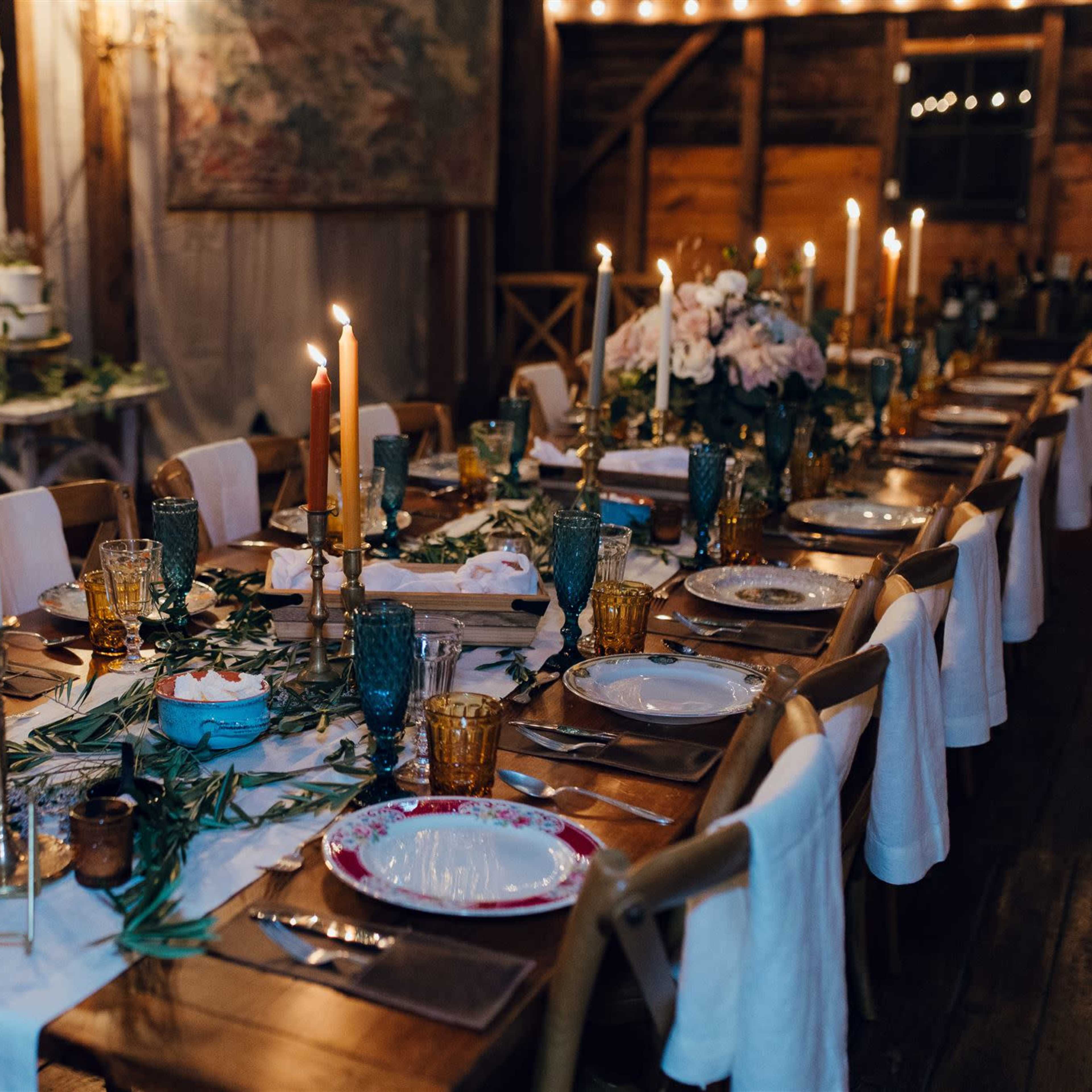 A long dining table is elegantly set with candles, glassware, and floral arrangements in a rustic indoor space.