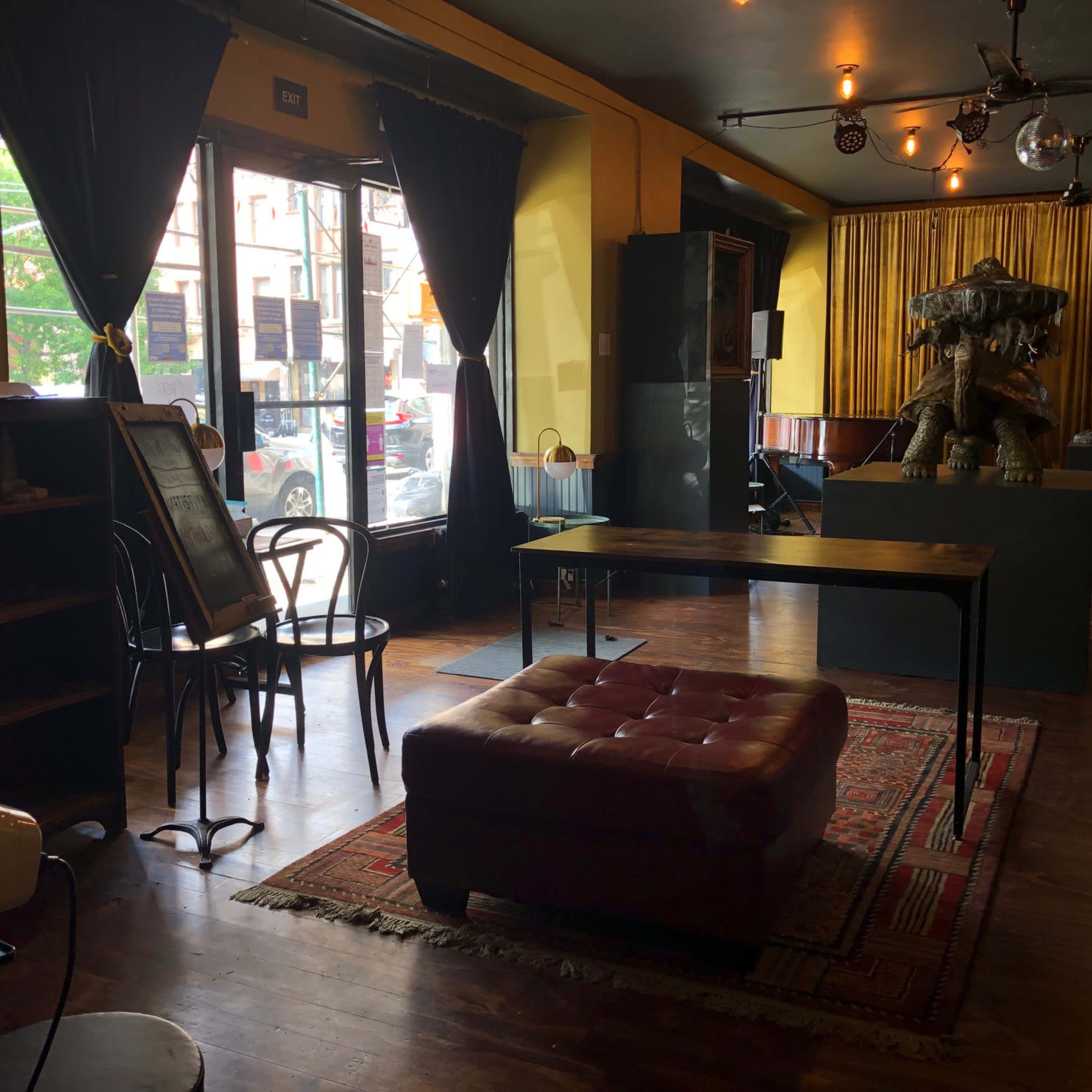 The interior of a small café features a wooden table, a plush ottoman, and a sculpture displayed on a raised platform, with large windows illuminating the space.