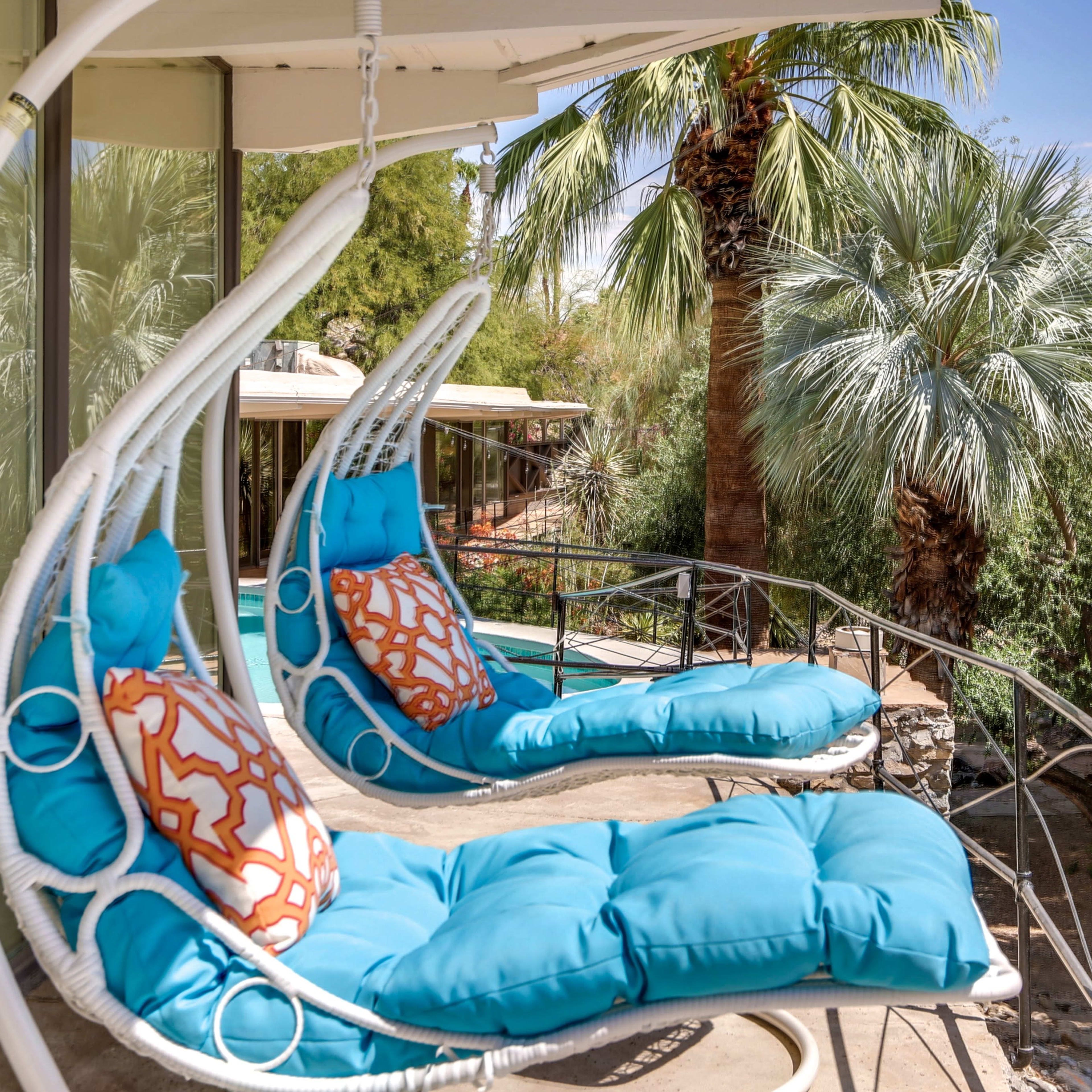 Two turquoise hanging chairs with decorative pillows are positioned on a balcony surrounded by palm trees.