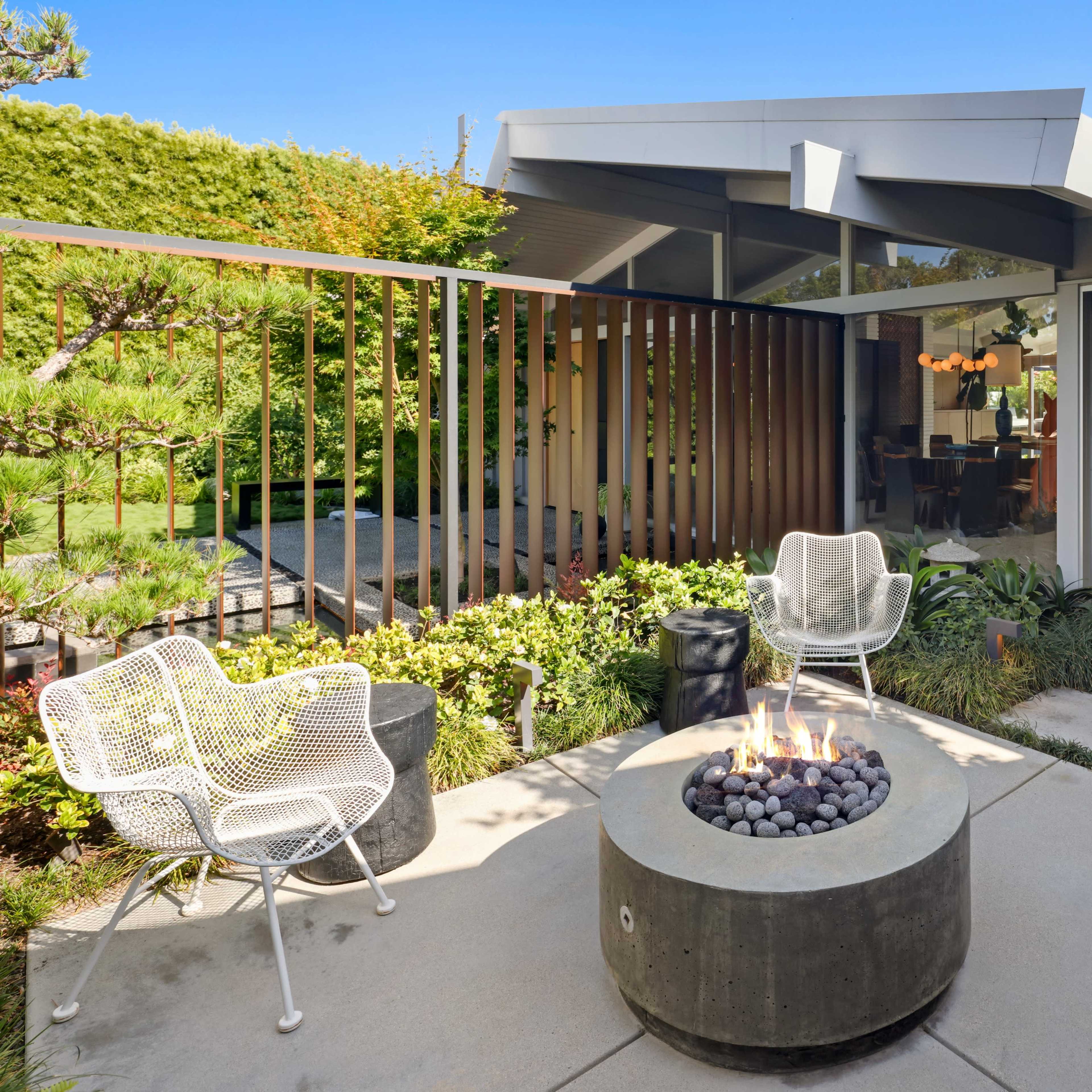 A modern outdoor seating area with four wire chairs arranged around a circular concrete fire pit, surrounded by greenery and leading to a contemporary home.