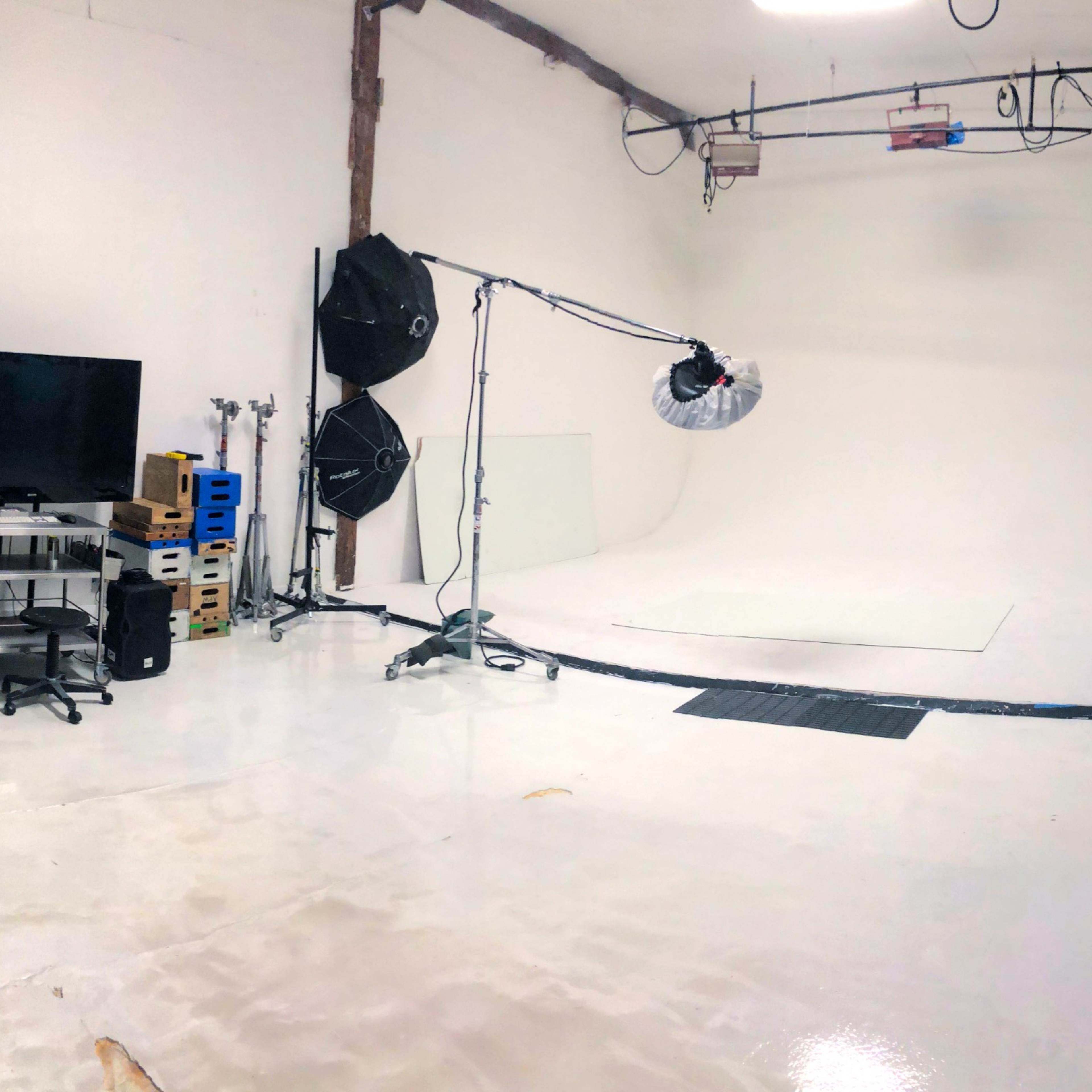 A spacious photography studio with a white backdrop, lighting equipment, a television, storage carts, and a white couch in the corner.