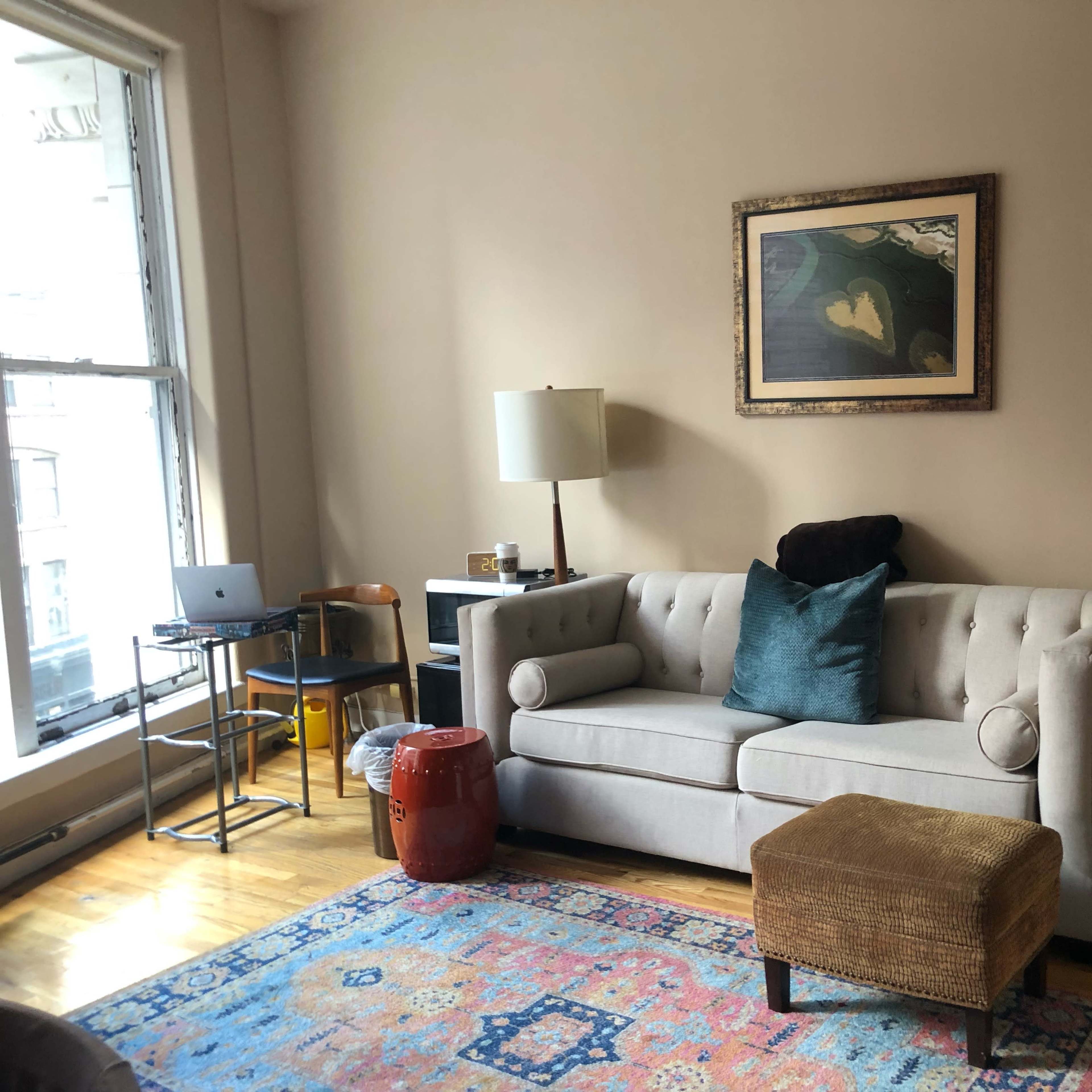 A living room with a gray tufted sofa, a small ottoman, a colorful area rug, and a desk with a laptop near a window.