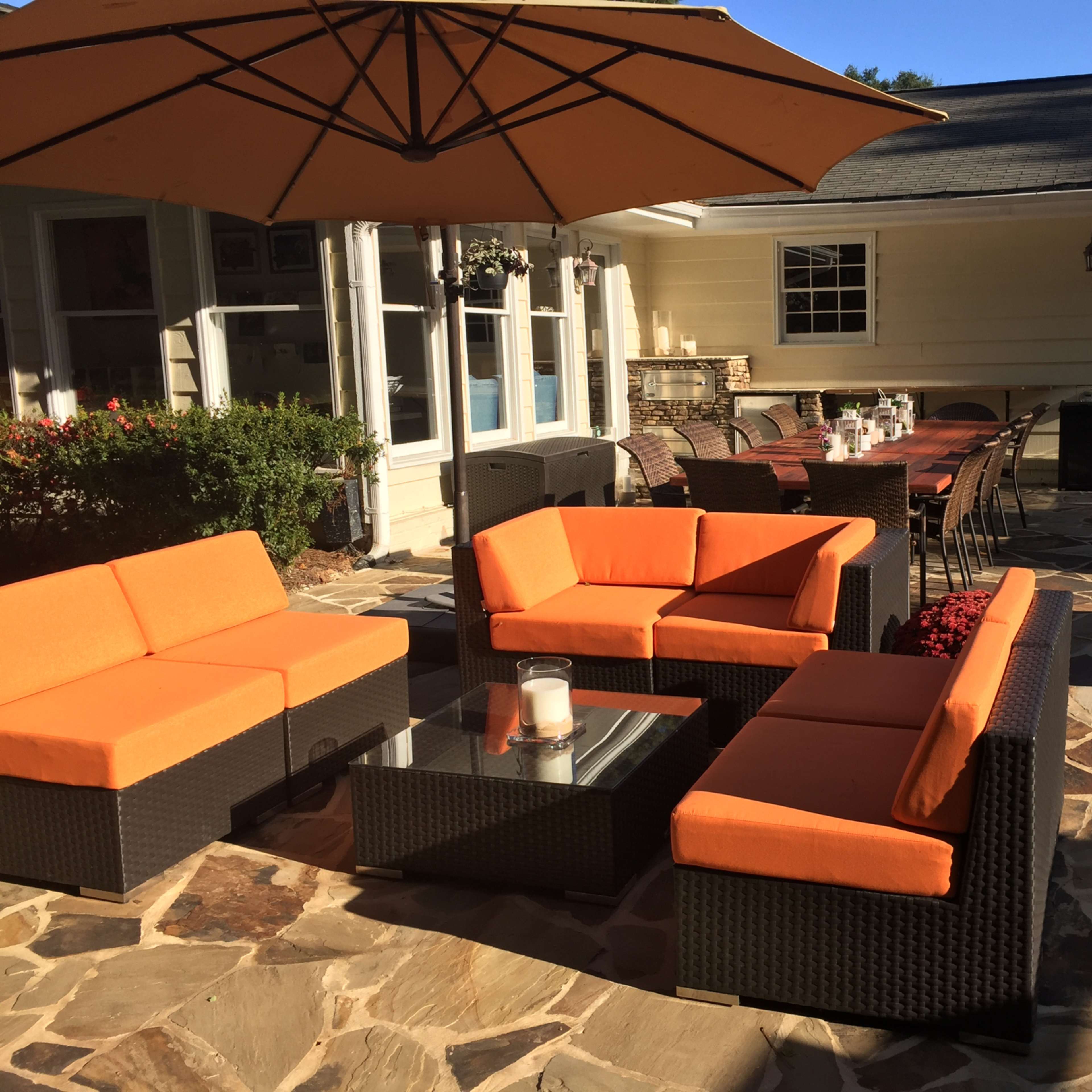 An outdoor seating area with orange cushions on wicker furniture, a large umbrella, and a stone patio adjacent to a house.