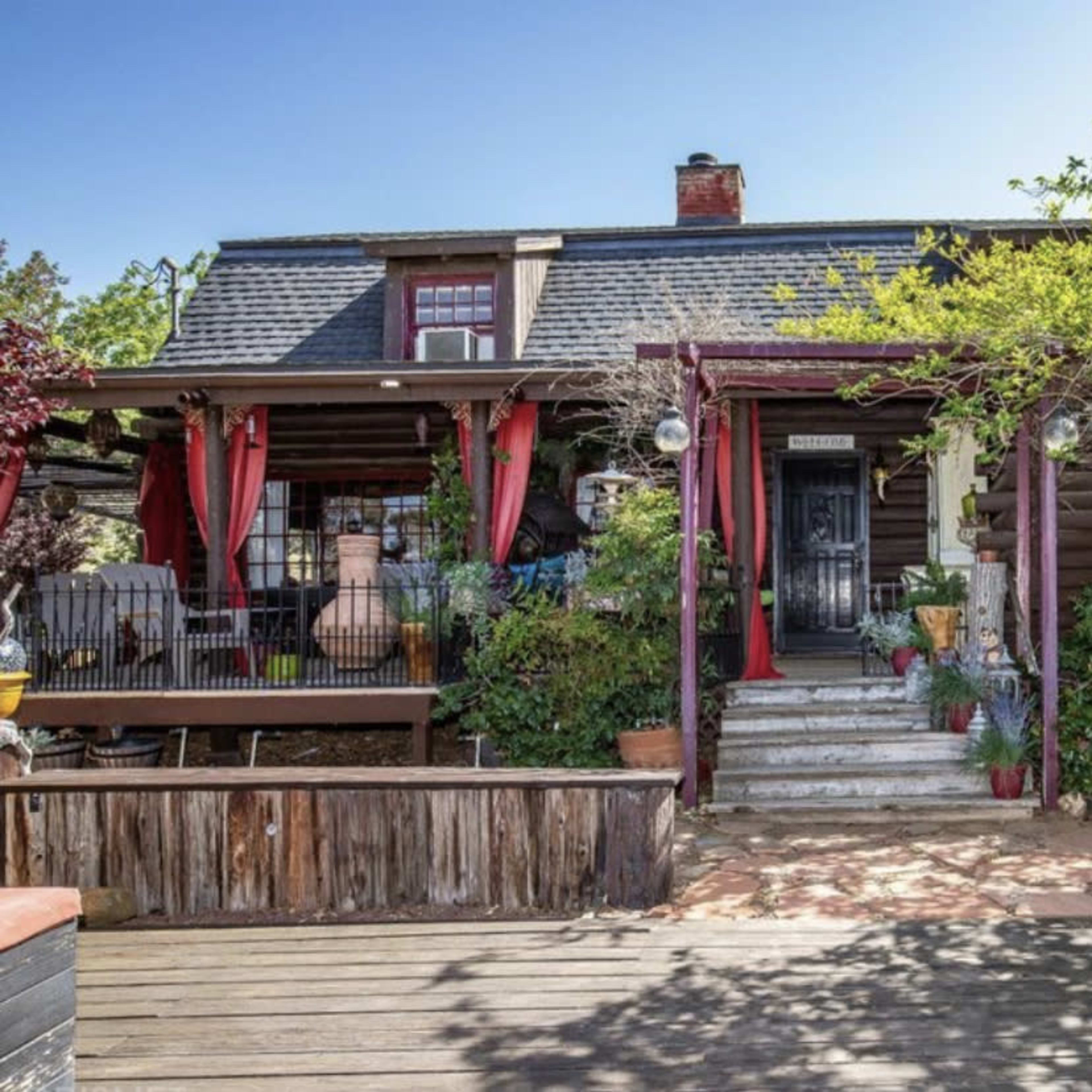 A rustic wooden house with a covered porch features greenery and colorful planters, leading up to a set of stairs.