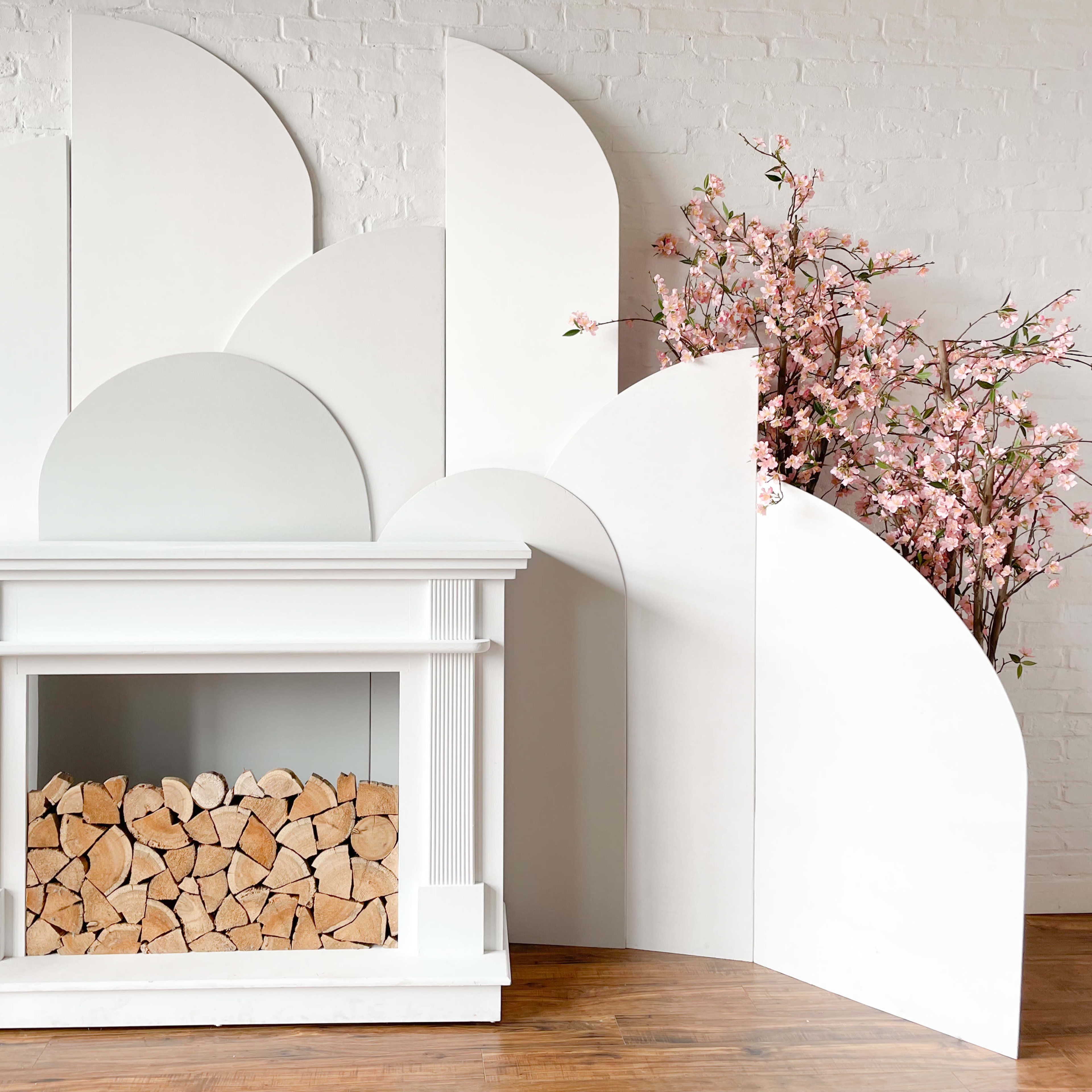 A white fireplace is set against a textured white wall, surrounded by large, curved white panels, with a decorative arrangement of pink cherry blossoms beside it.