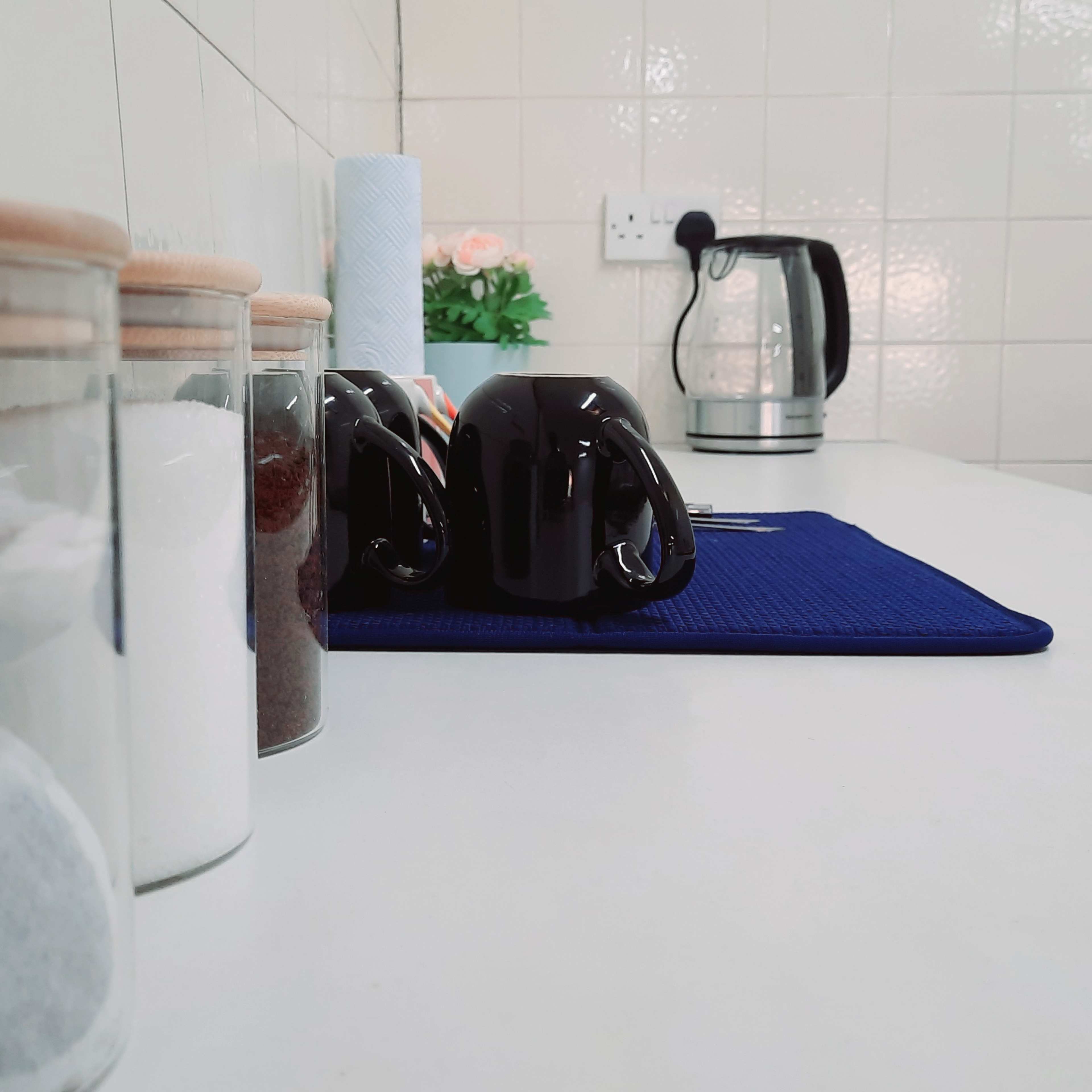 A kitchen countertop with glass containers, black coffee mugs, a kettle, and a roll of paper towels.
