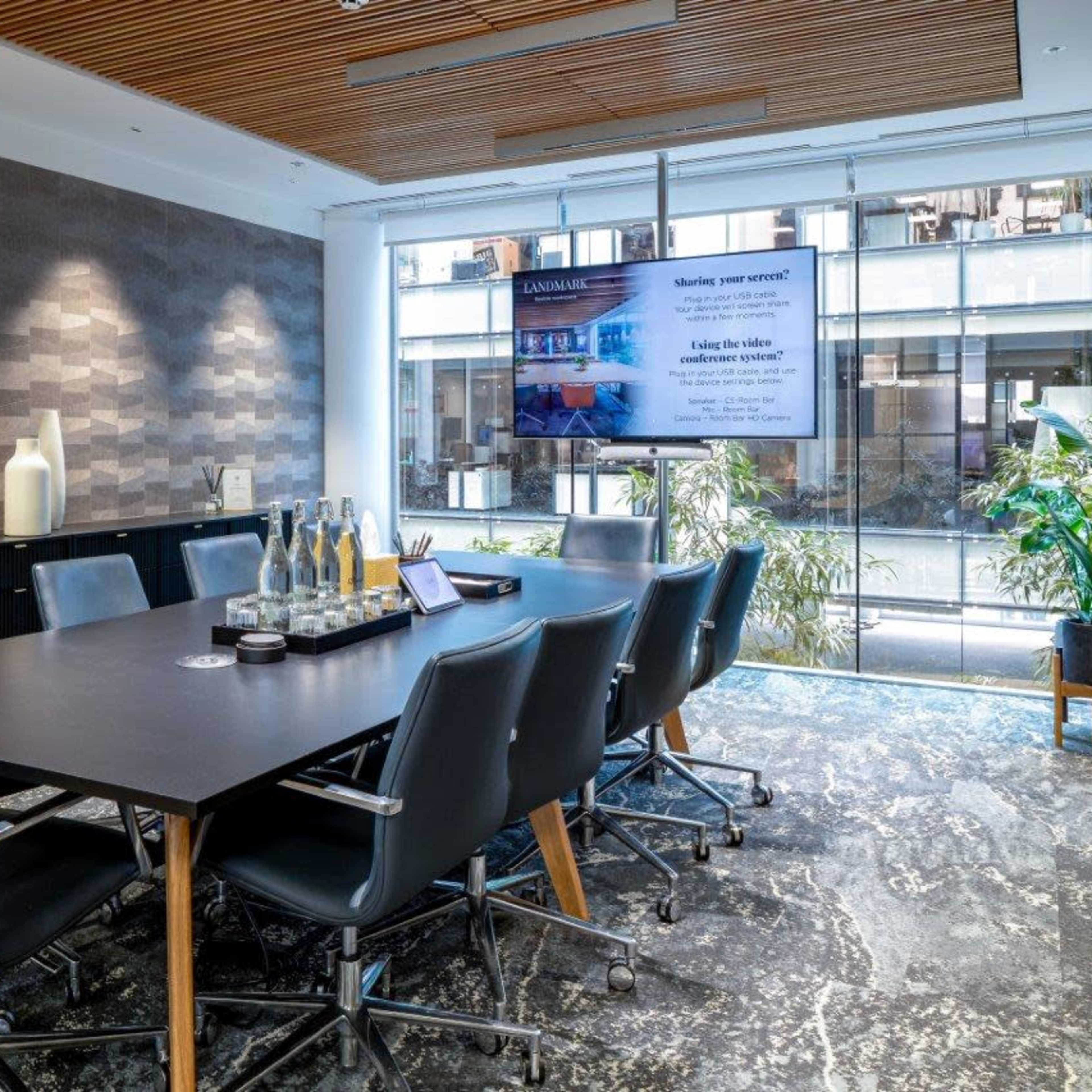 A modern conference room features a large table with ergonomic chairs, a television screen displaying a presentation, and plants visible through large windows.