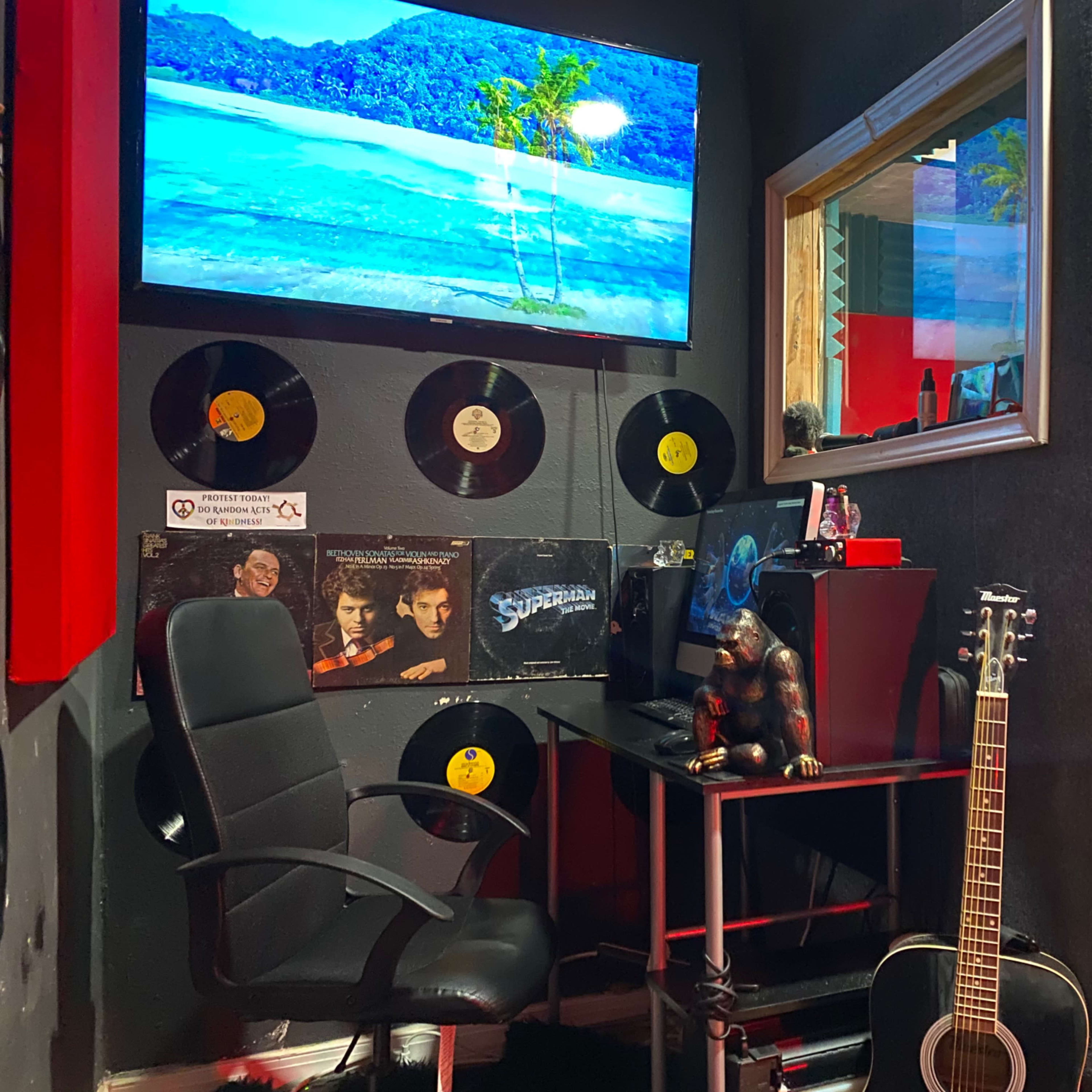 A music studio corner with a black chair, a desk holding a computer and a sculpture, a guitar leaning against the wall, and records displayed alongside a TV screen showing a beach scene.