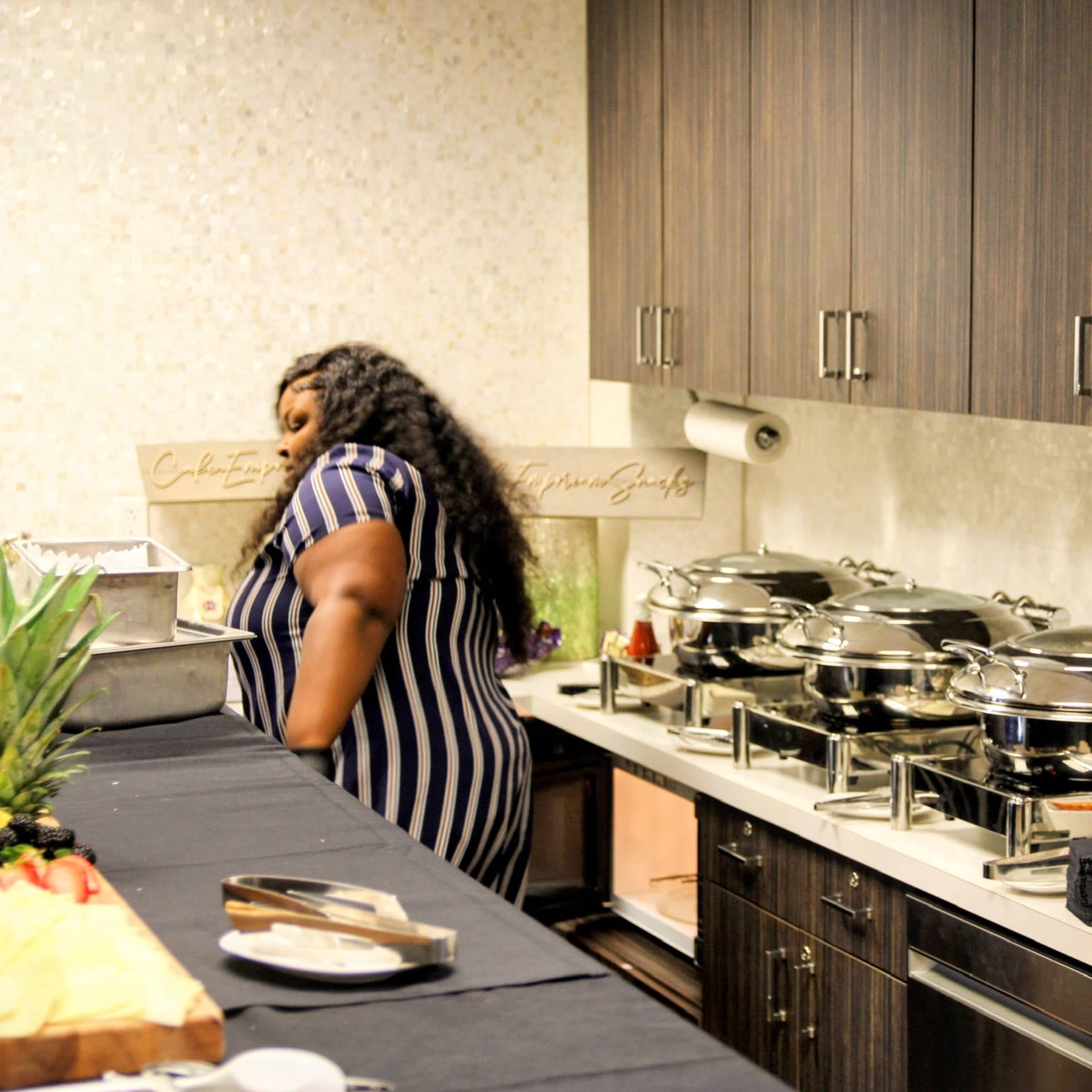 A person in a striped dress stands near a buffet table with three large chafing dishes and stacked plates in a modern kitchen.