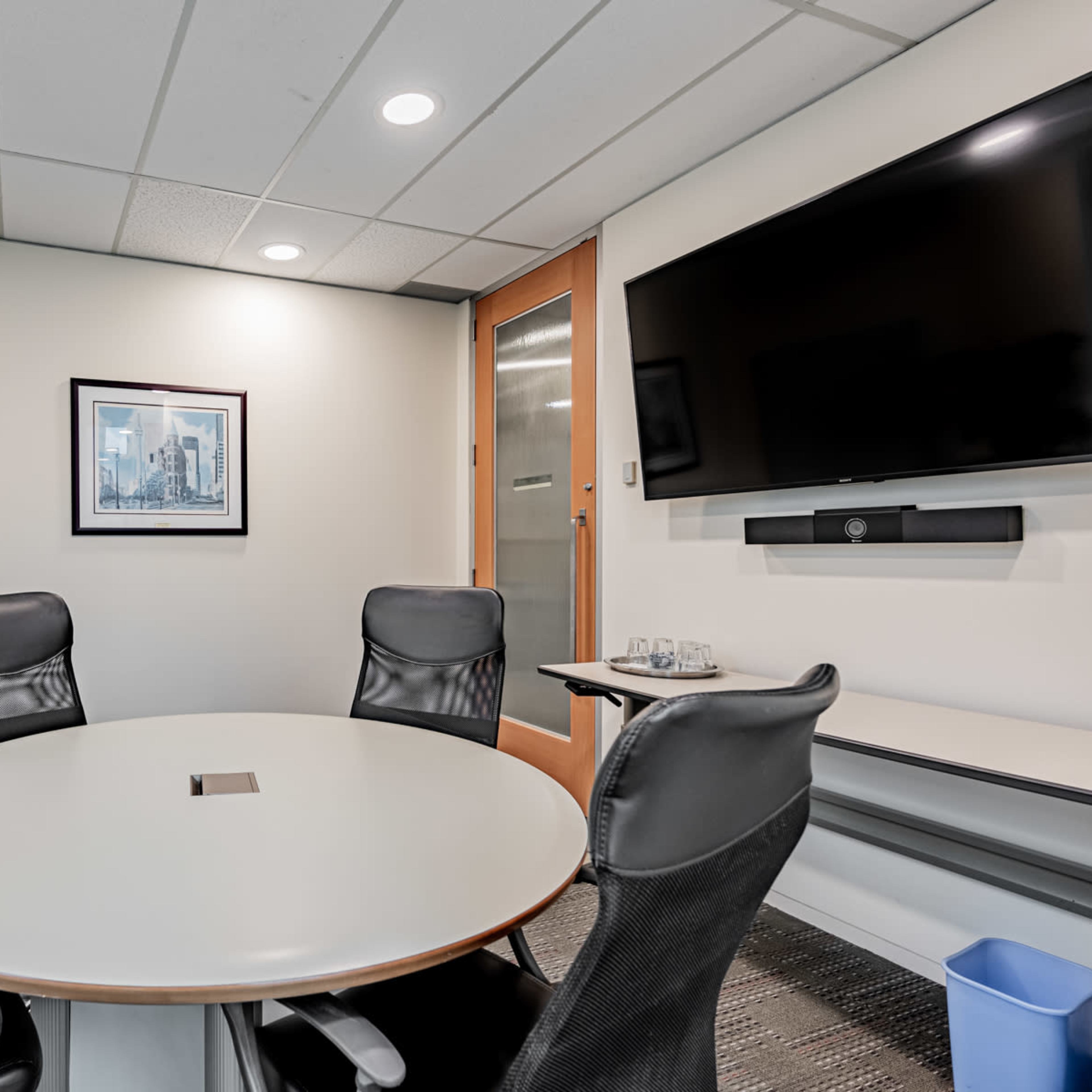 A modern conference room features a round table surrounded by black ergonomic chairs, a large wall-mounted television, and a glass door.