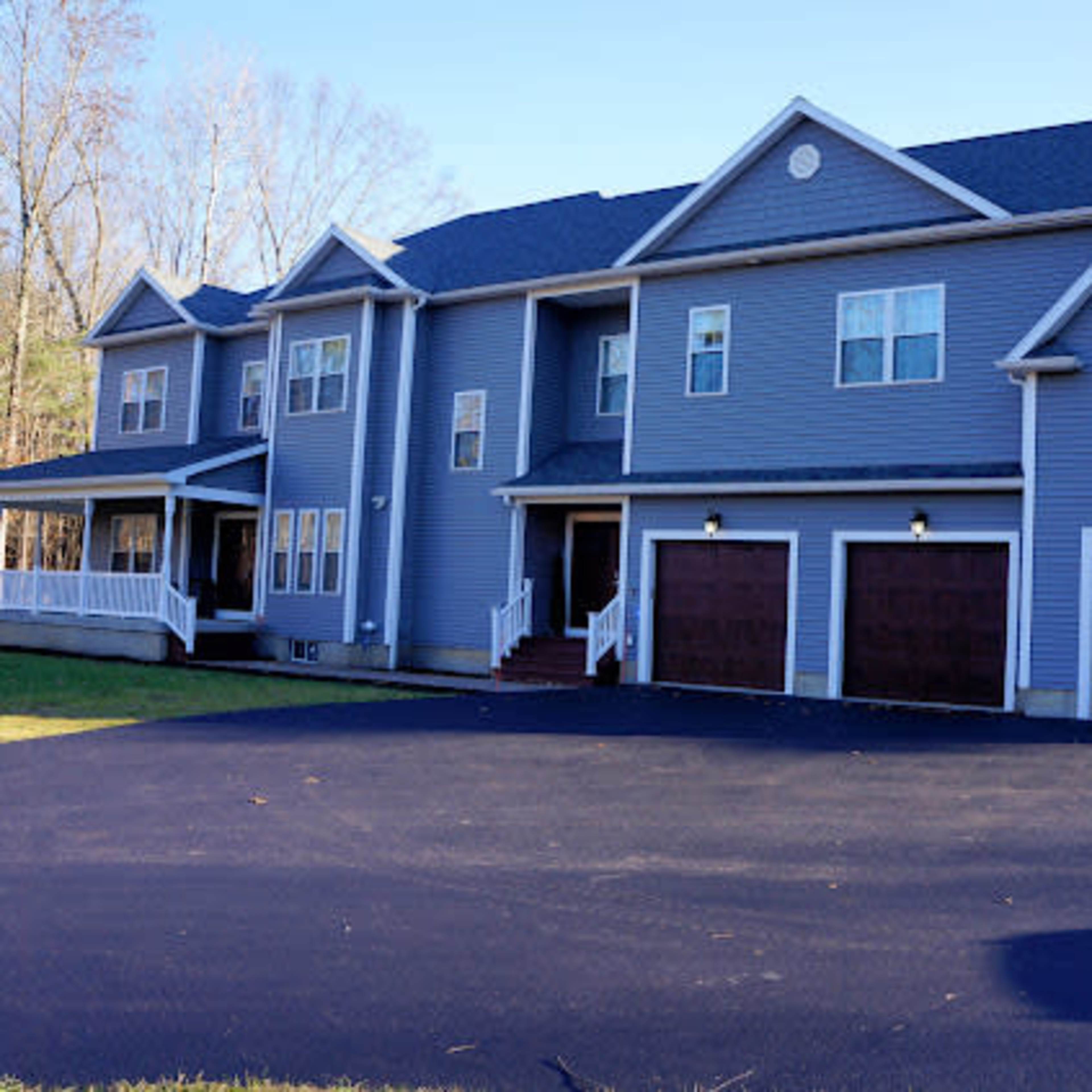 A two-story blue house with a three-car garage is set on a paved driveway surrounded by trees.