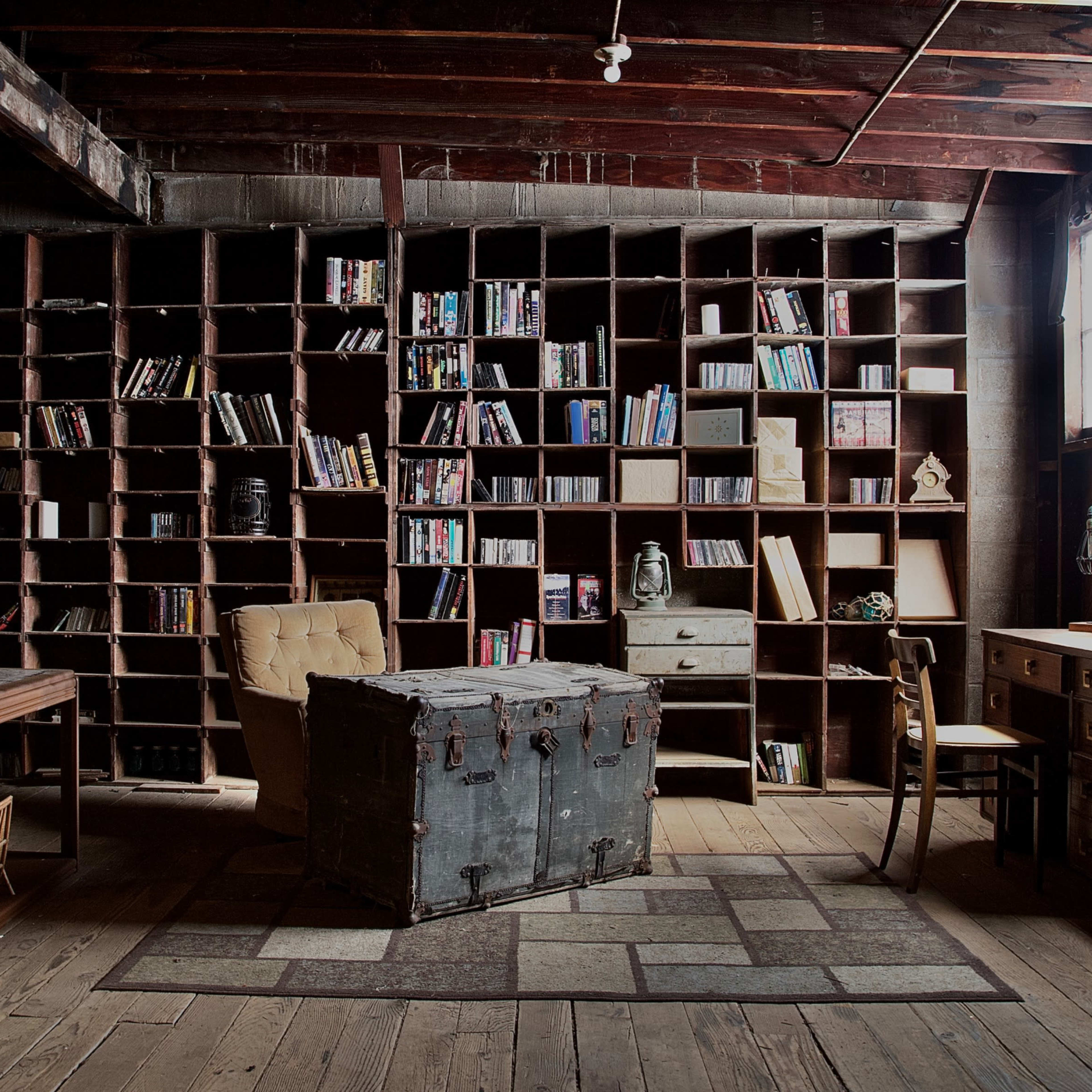 A cozy library with wooden shelving filled with books and a vintage trunk in the center of the room alongside a desk and chairs.